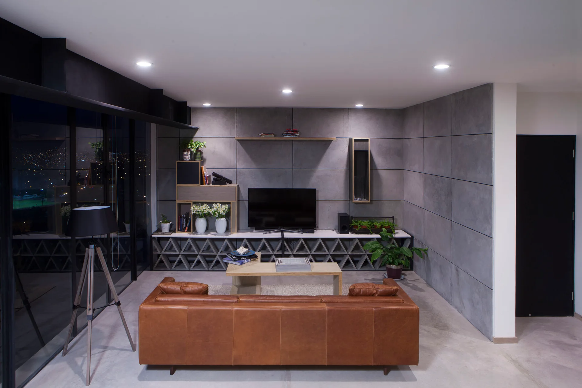 A modern living room features a tan leather sofa, a geometric concrete media console, and grey grid wall paneling.