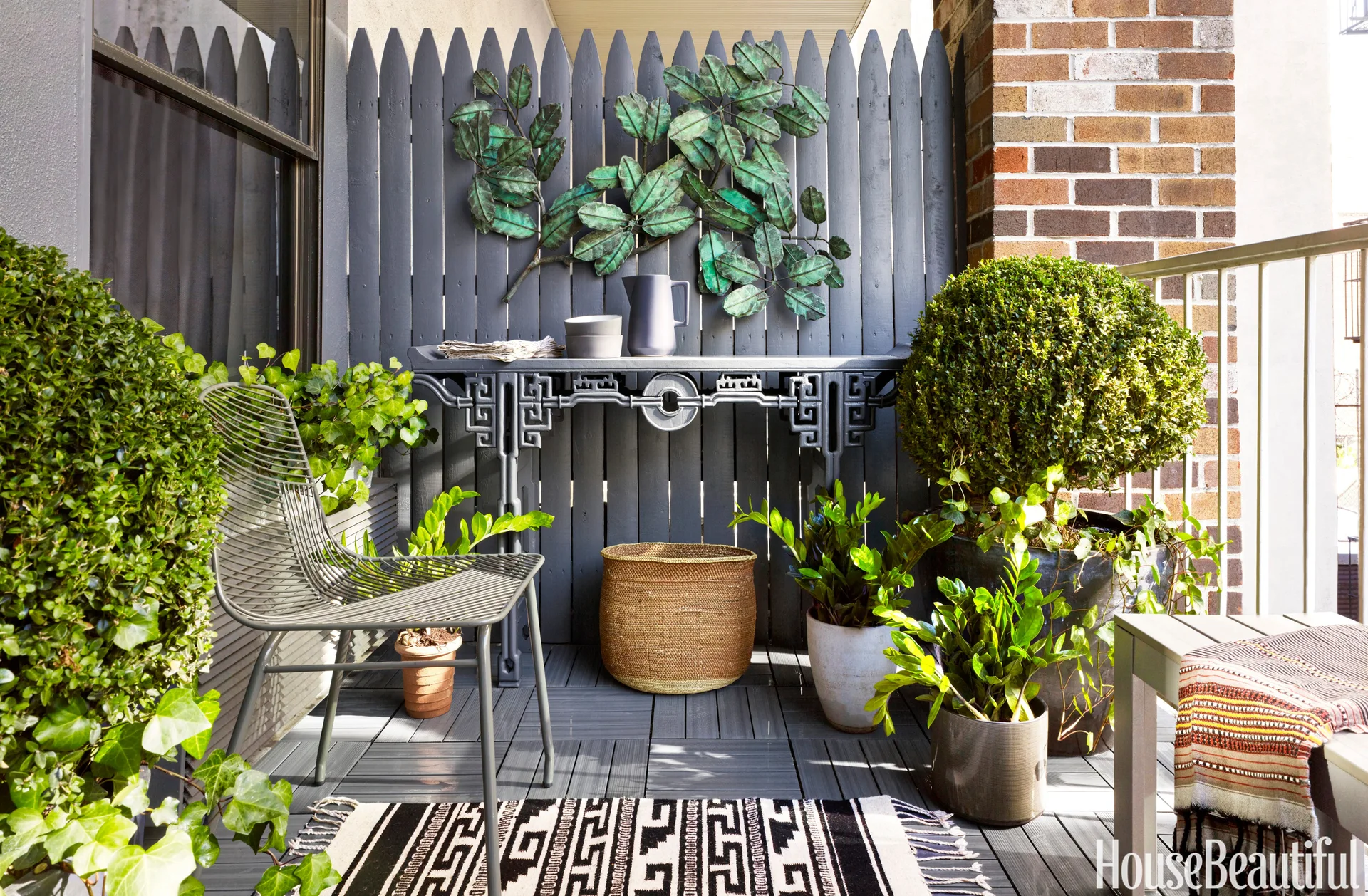 A small balcony features a grey painted picket fence decorated with a metal leaf sculpture, a matching grey console table, potted plants, and a wire chair.