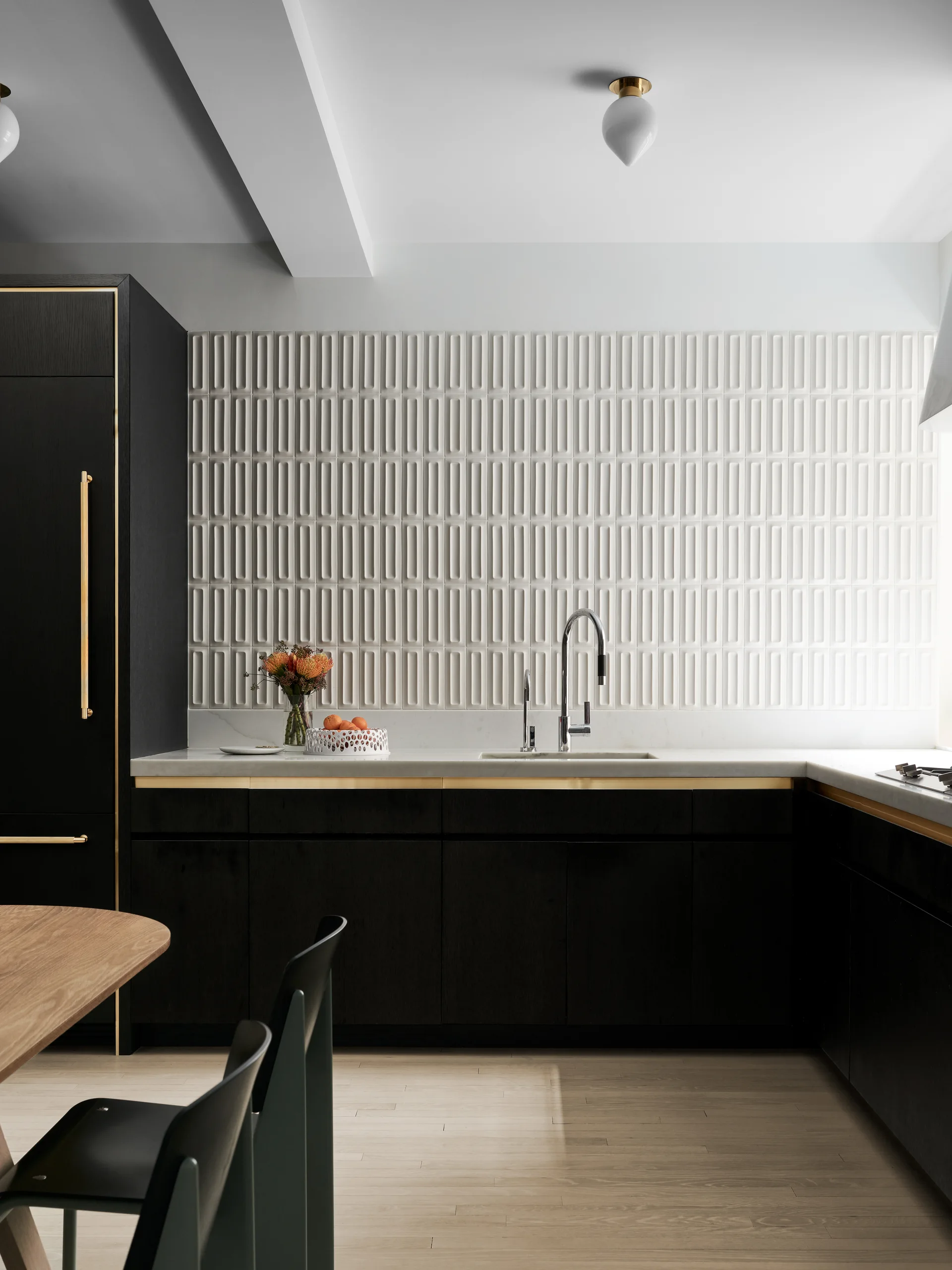 A modern kitchen featuring matte black cabinets with gold trim, a textured white tile backsplash, and light wood flooring.