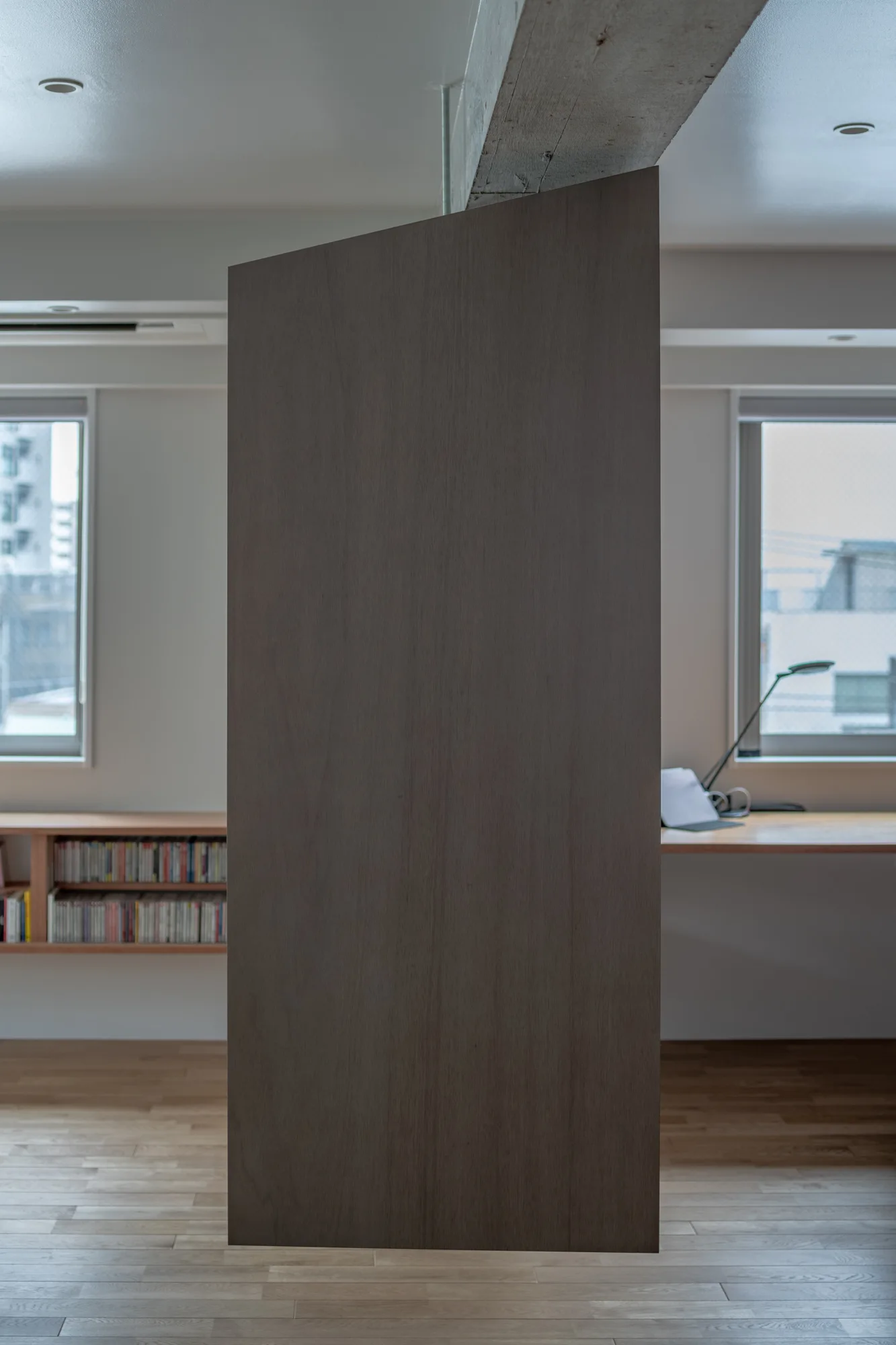 A central dark wood room partition stands between a built-in desk and low bookshelf under an exposed concrete ceiling beam in a minimalist workspace.