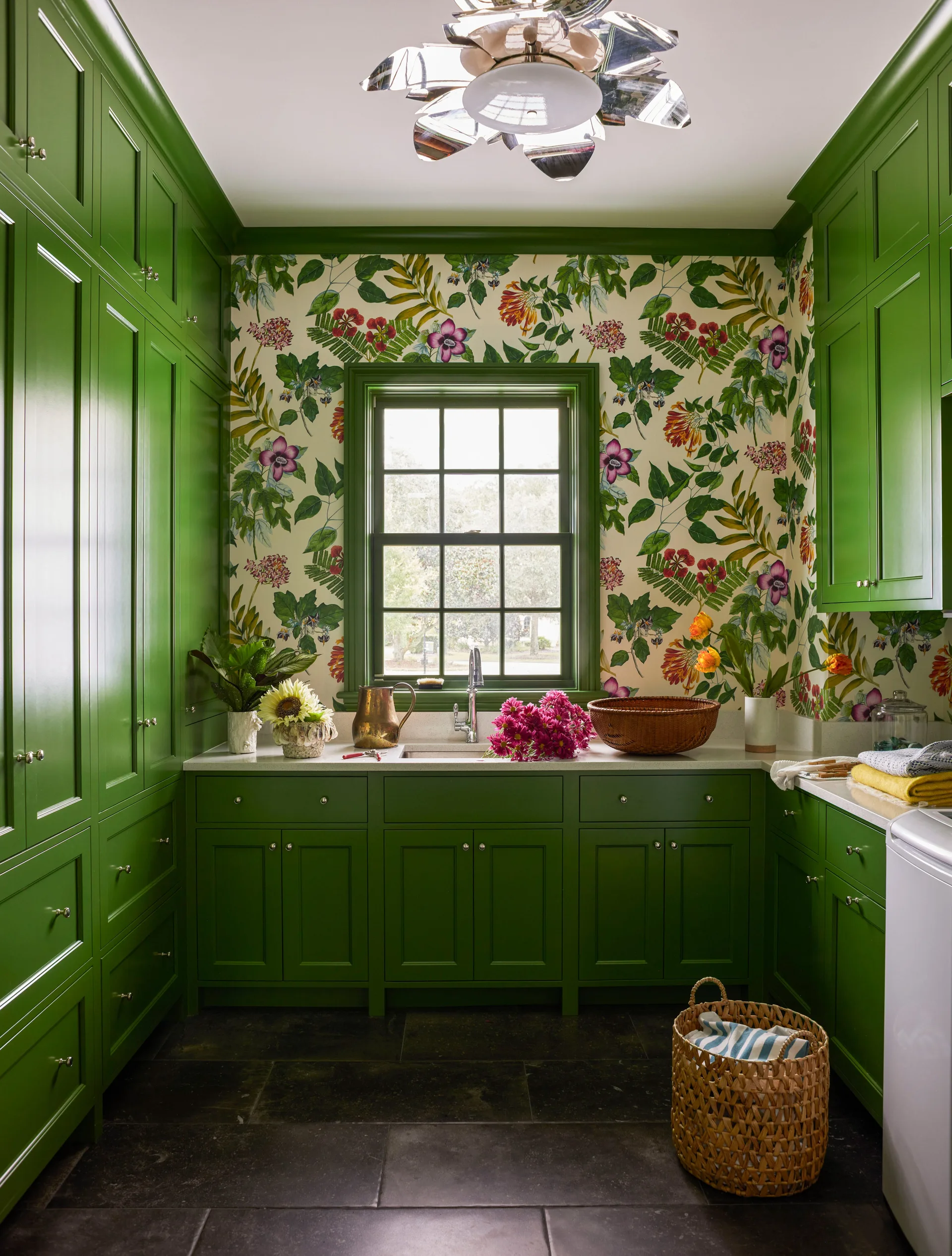 A laundry room features glossy kelly green cabinets, botanical floral wallpaper, a central window over a utility sink, and dark stone tile flooring.