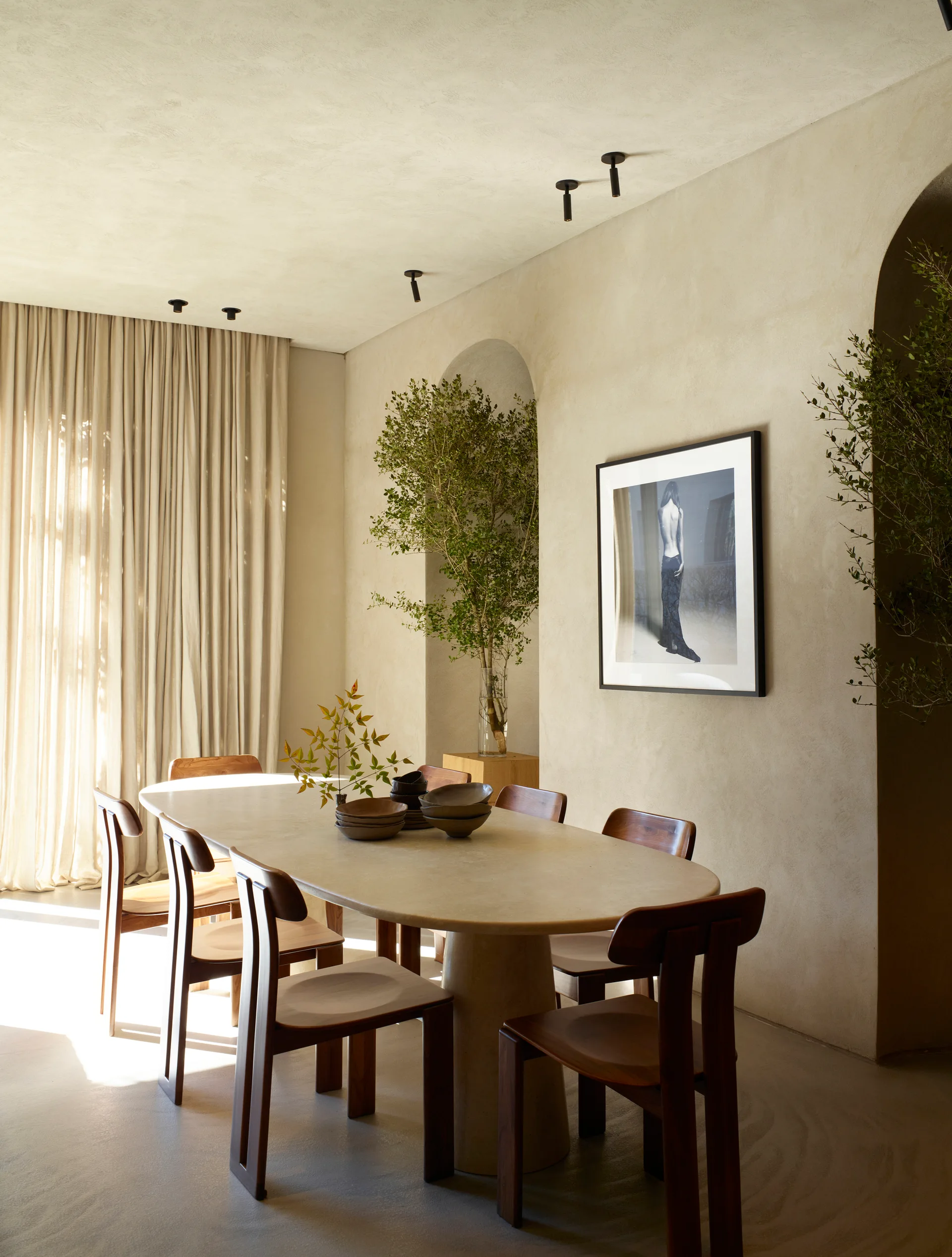 A minimalist dining room features a large oval stone table surrounded by wooden chairs set against beige plaster walls with arched niches.