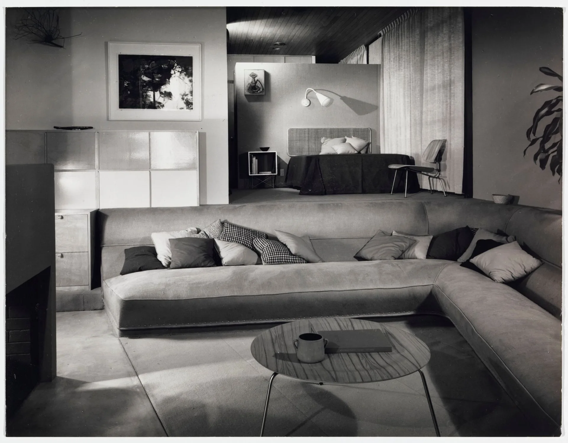 Black and white photo of a mid-century modern sunken living room with a large sectional sofa, a low wood coffee table, and an elevated bedroom area.