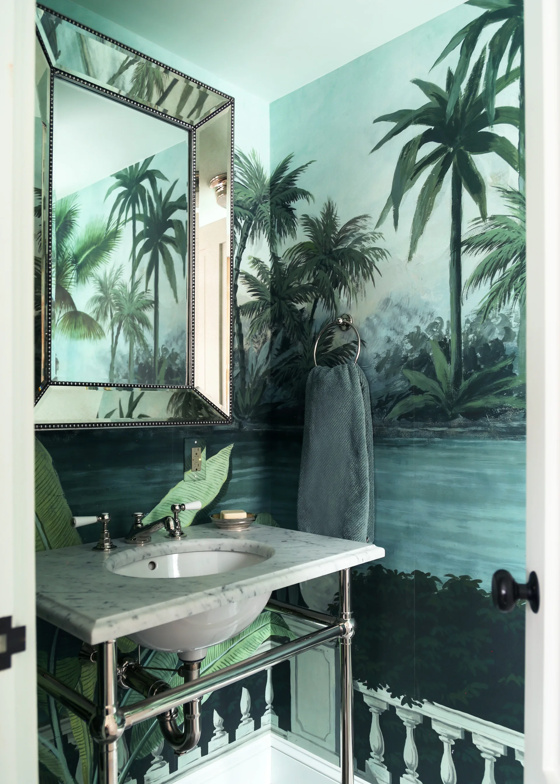 A compact powder room with tropical palm tree mural wallpaper, a marble console sink with chrome legs, and a beveled wall mirror.