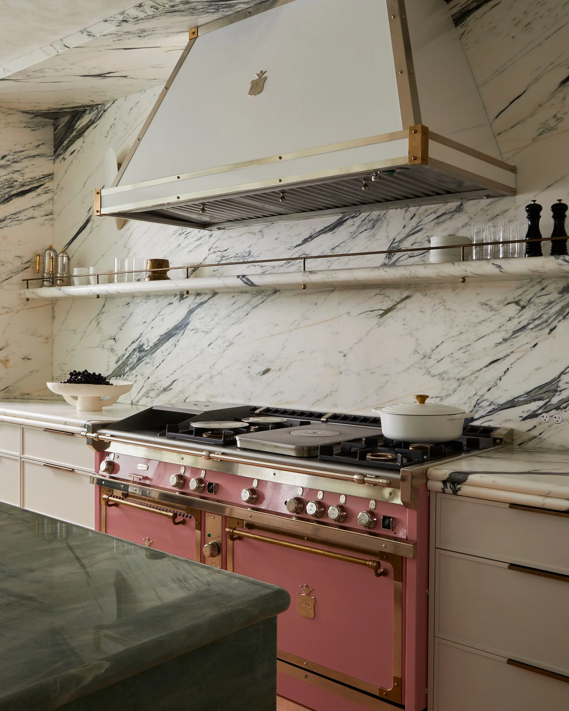 A pink kitchen range sits beneath a white and brass hood against a white marble backsplash with heavy black veining and a matching floating shelf.