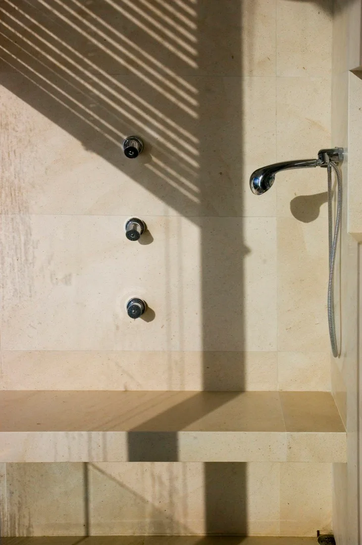 A close-up view of a shower interior showing large-format beige stone tiles, an integrated stone bench, and chrome hand-held shower fixtures.