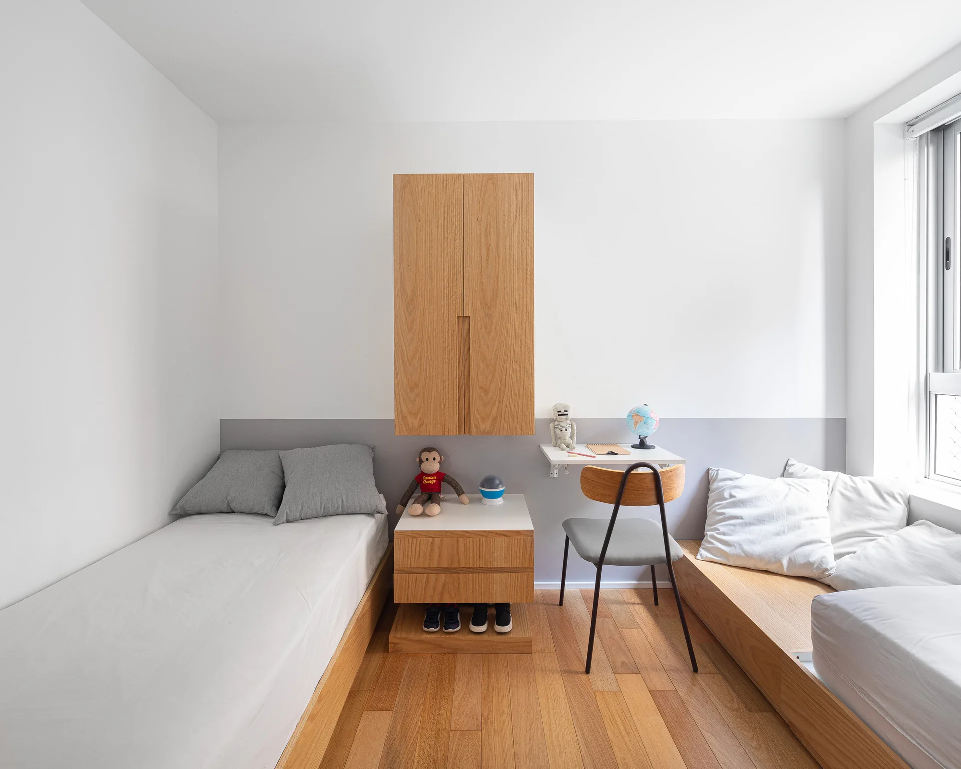 A minimalist children's bedroom featuring two wooden platform beds, a floating desk, and a wall-mounted wooden cabinet against a white and grey wall.