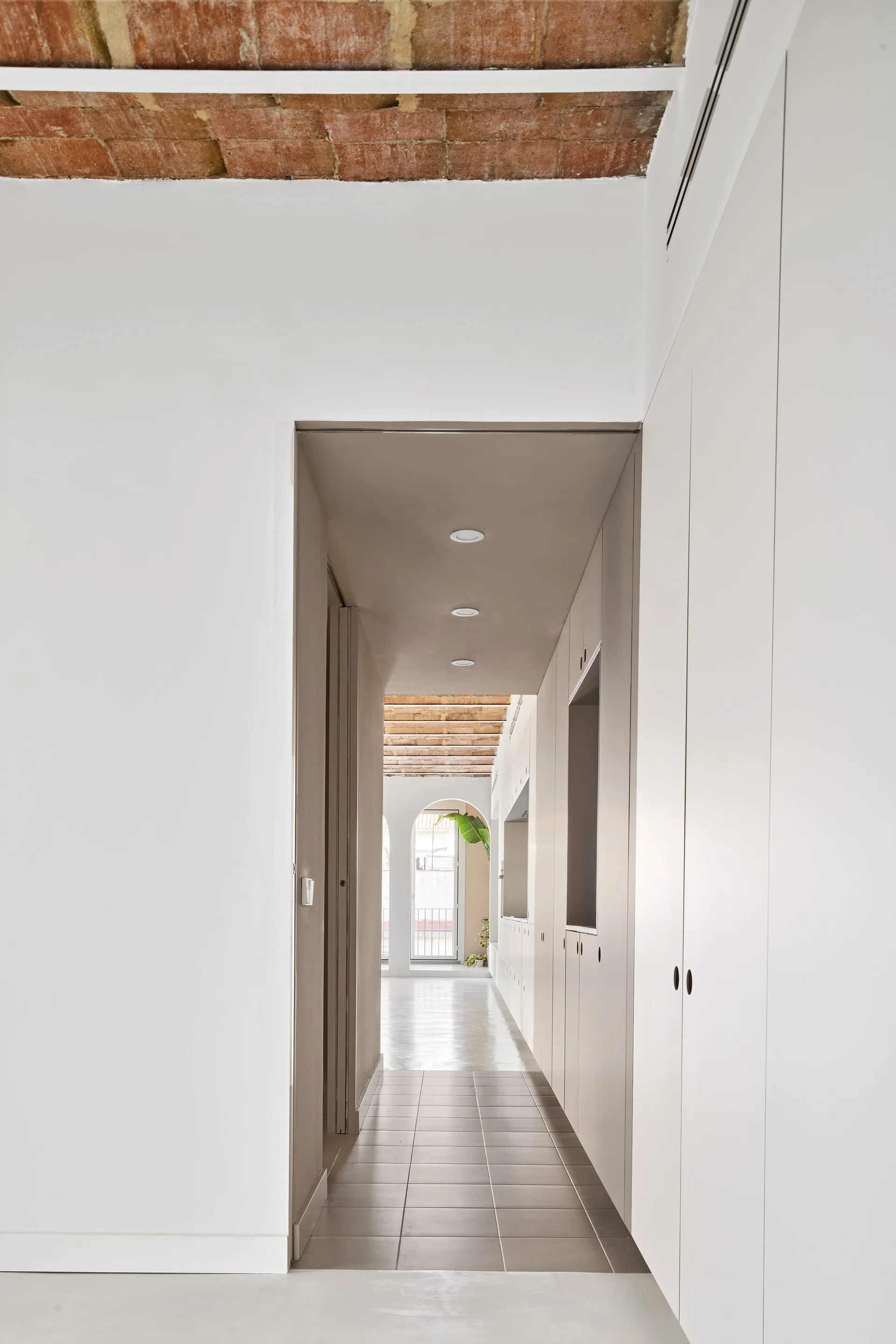 A minimalist hallway featuring white built-in storage cabinets, grey tile floors, and a vaulted ceiling with exposed brick.