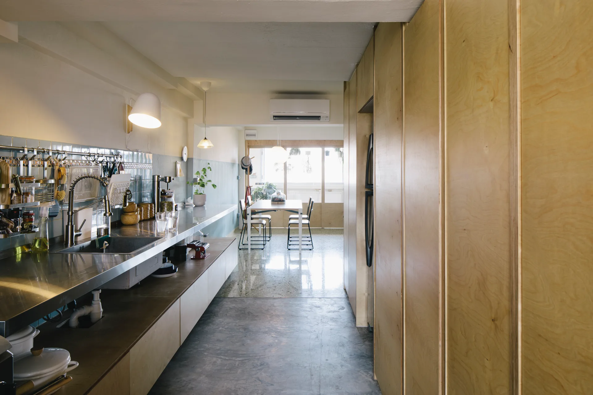 A long galley kitchen features full-height plywood cabinets on one side and a stainless steel countertop with a blue tile backsplash on the other.
