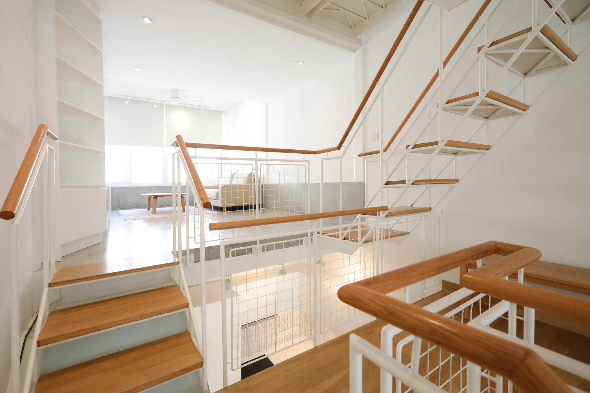 A white metal staircase with light wood treads and grid railings connects different levels of a bright, minimalist apartment.
