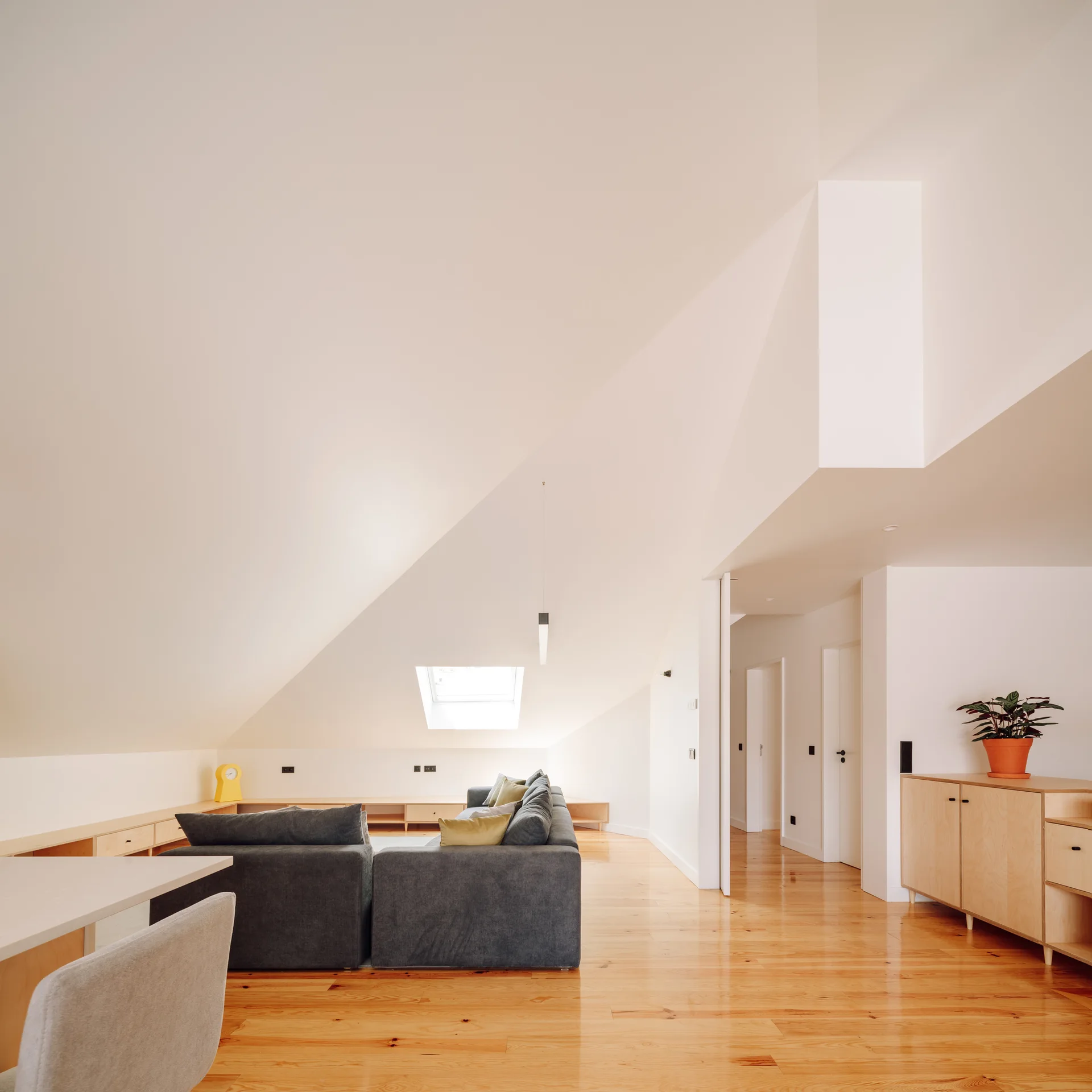 A minimalist attic living room features a dark grey sectional sofa on glossy wood floors beneath a dramatic sloped white ceiling with a skylight.