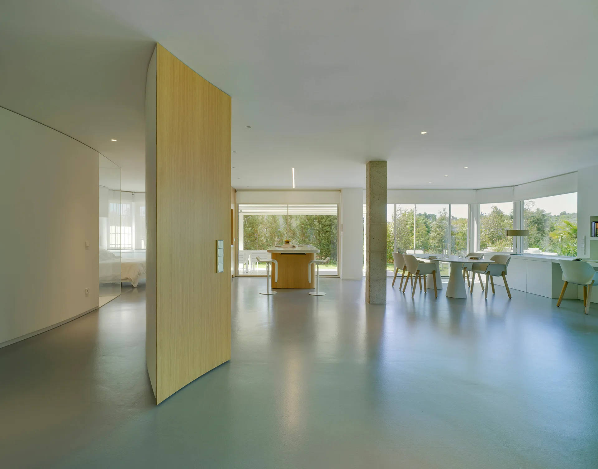 A minimalist open-concept living space with a large blonde wood pivoting door, seamless gray flooring, a stone column, and floor-to-ceiling windows.