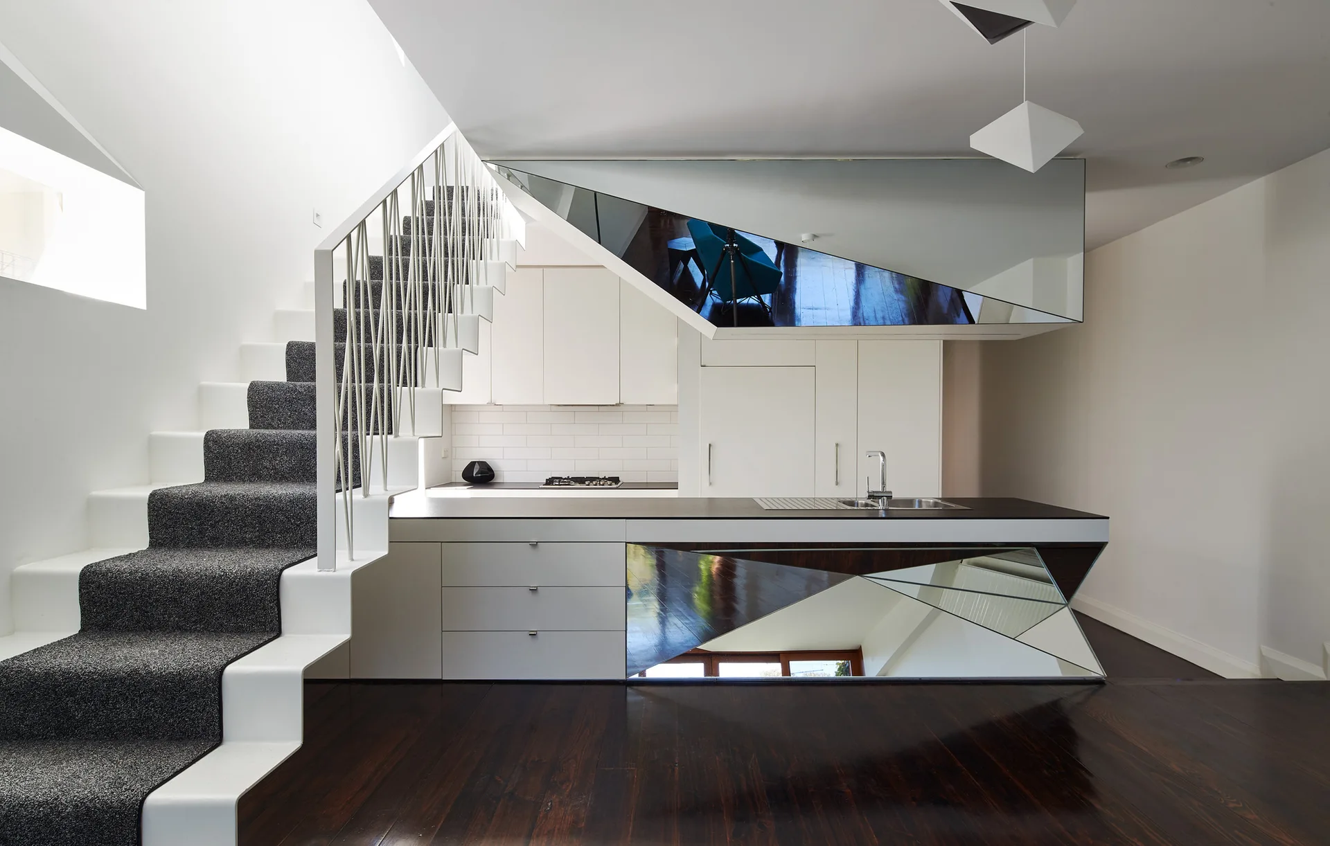 A modern white kitchen features a dark countertop island with a geometric mirrored panel next to a white staircase with a grey carpet runner.