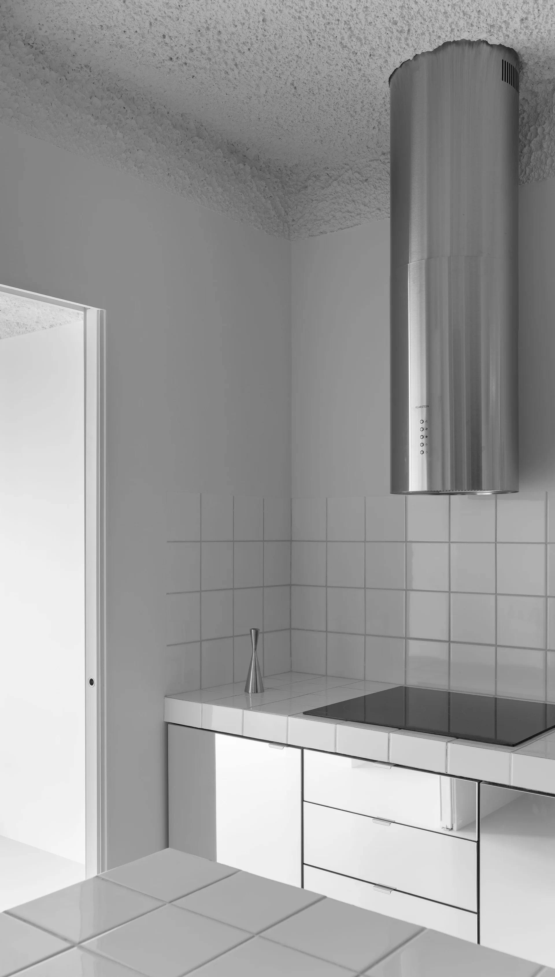 A minimalist kitchen featuring a cylindrical stainless steel range hood, white square tiled countertops and backsplash, and flat-front white cabinets.