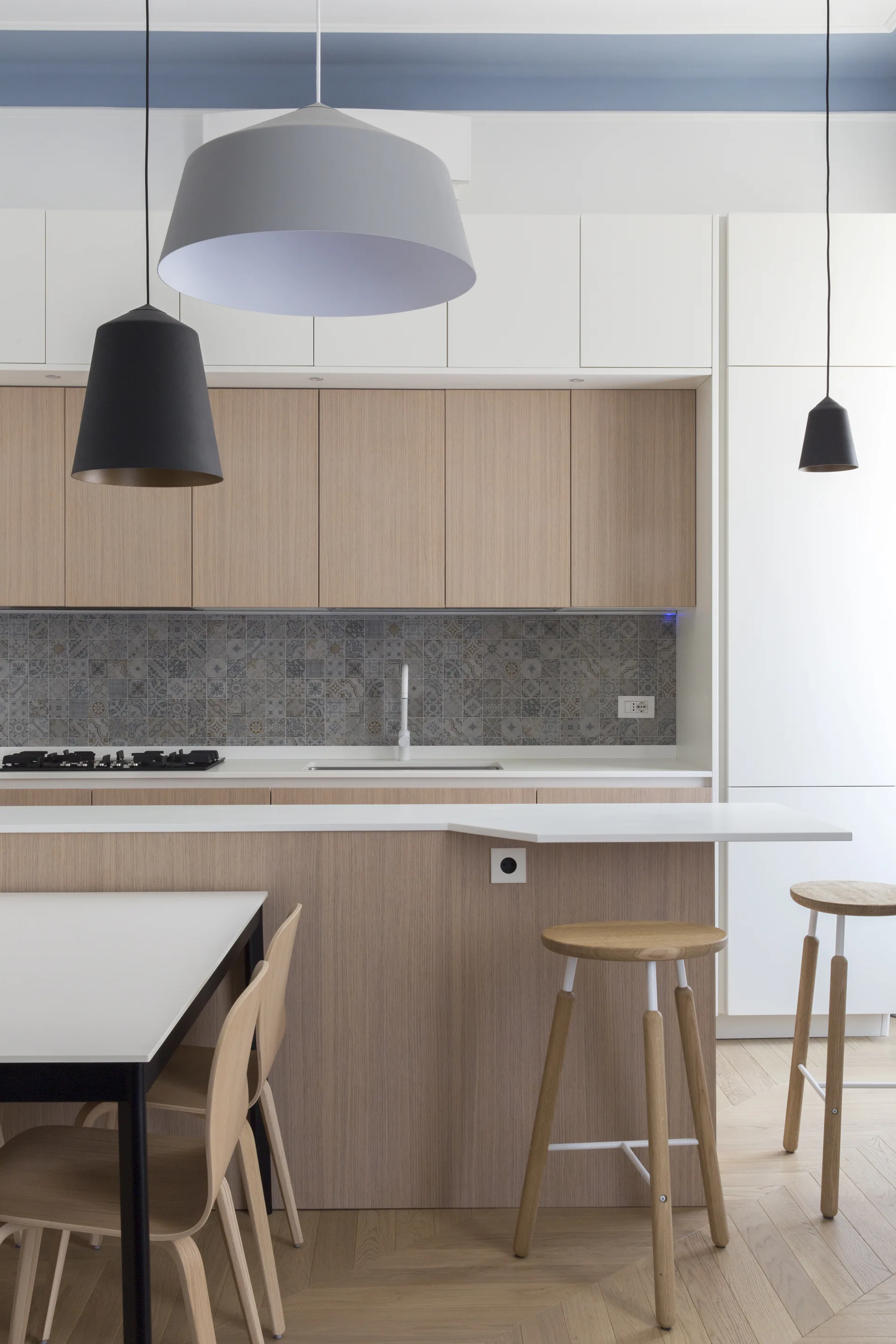 A modern kitchen featuring light wood and white cabinetry, a patterned tile backsplash, a kitchen island with bar stools, and herringbone wood floors.