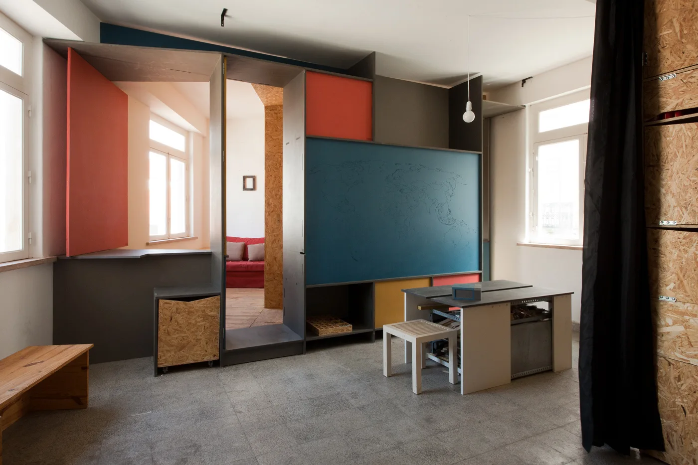 A modern home office features a large multi-colored partition wall with integrated shelving, a small desk, and a view of a red sofa in the background.
