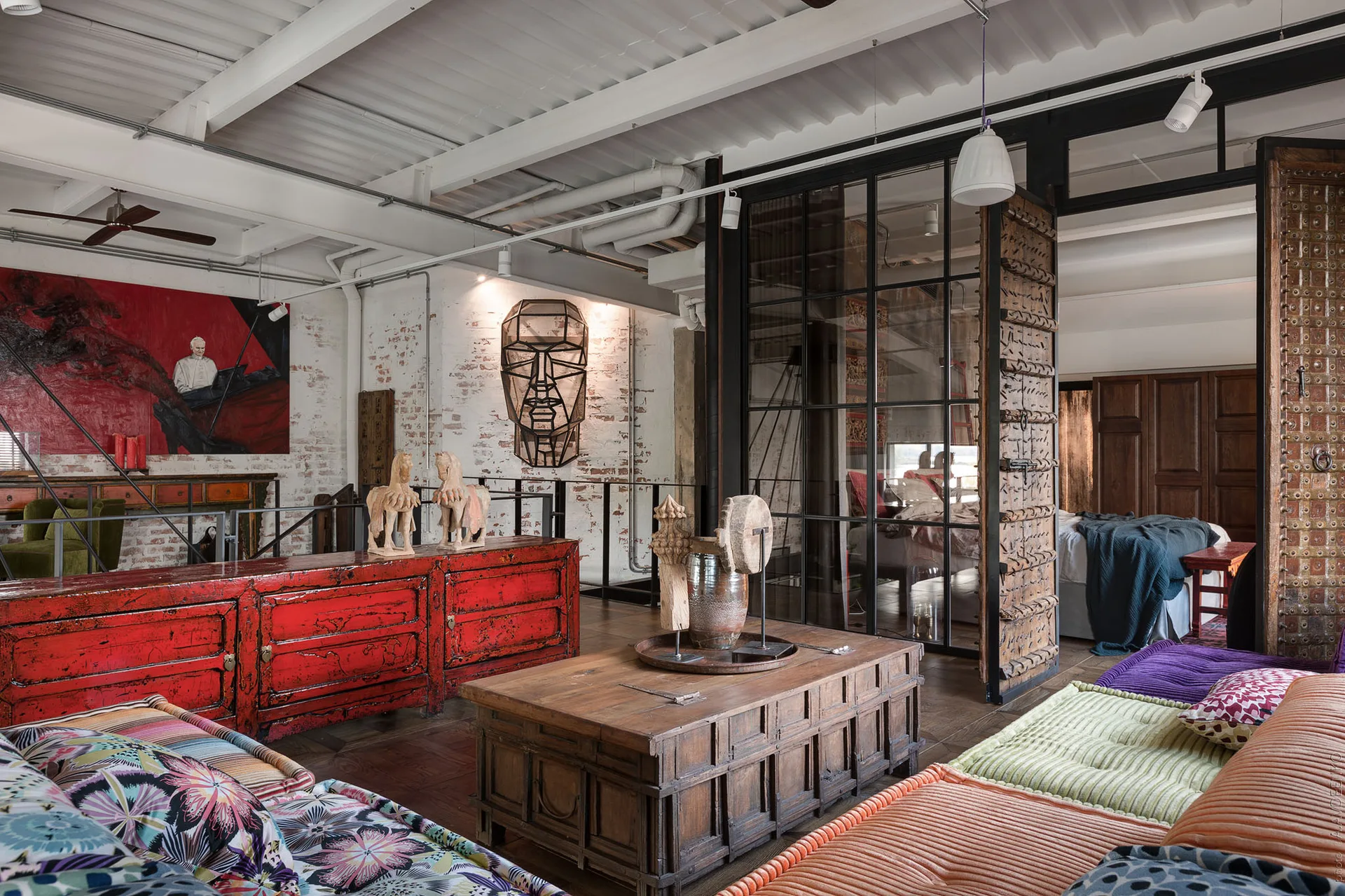 An eclectic industrial loft living room with white brick walls, a distressed red sideboard, a wooden trunk coffee table, and a glass partition.
