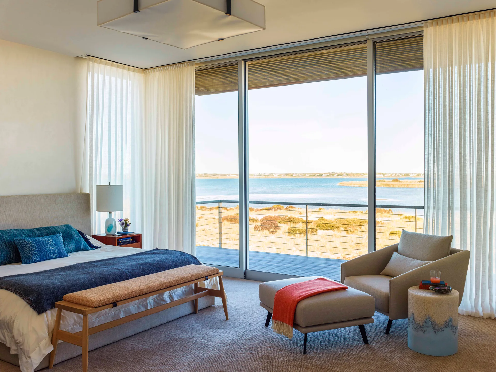 A coastal bedroom features a bed with blue accent pillows, an armchair with an ottoman, and floor-to-ceiling windows showing a water view.