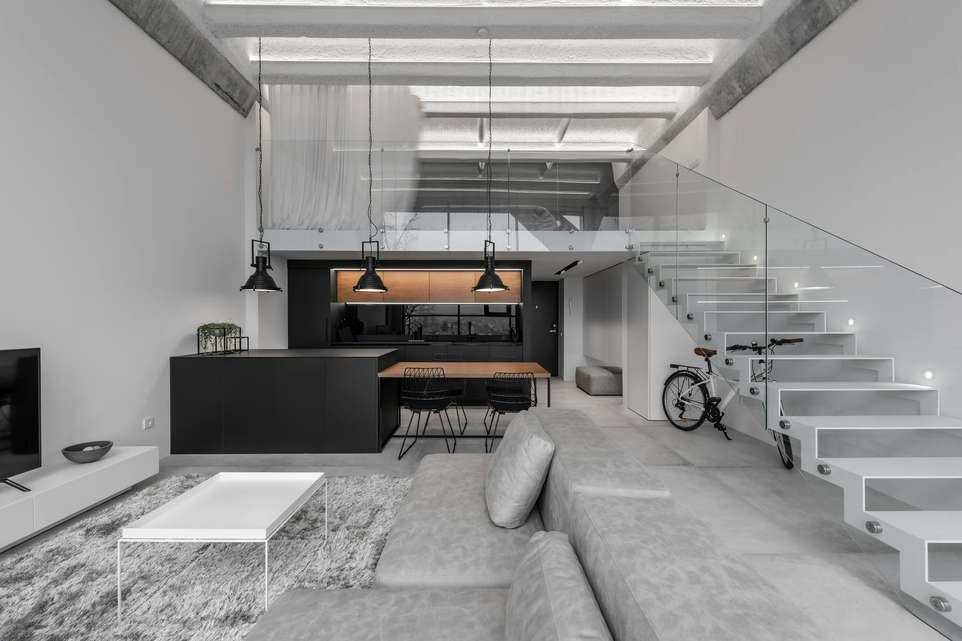 An open-plan loft with a grey sectional sofa, white floating staircase, glass railing, and black kitchen island under exposed concrete beams.