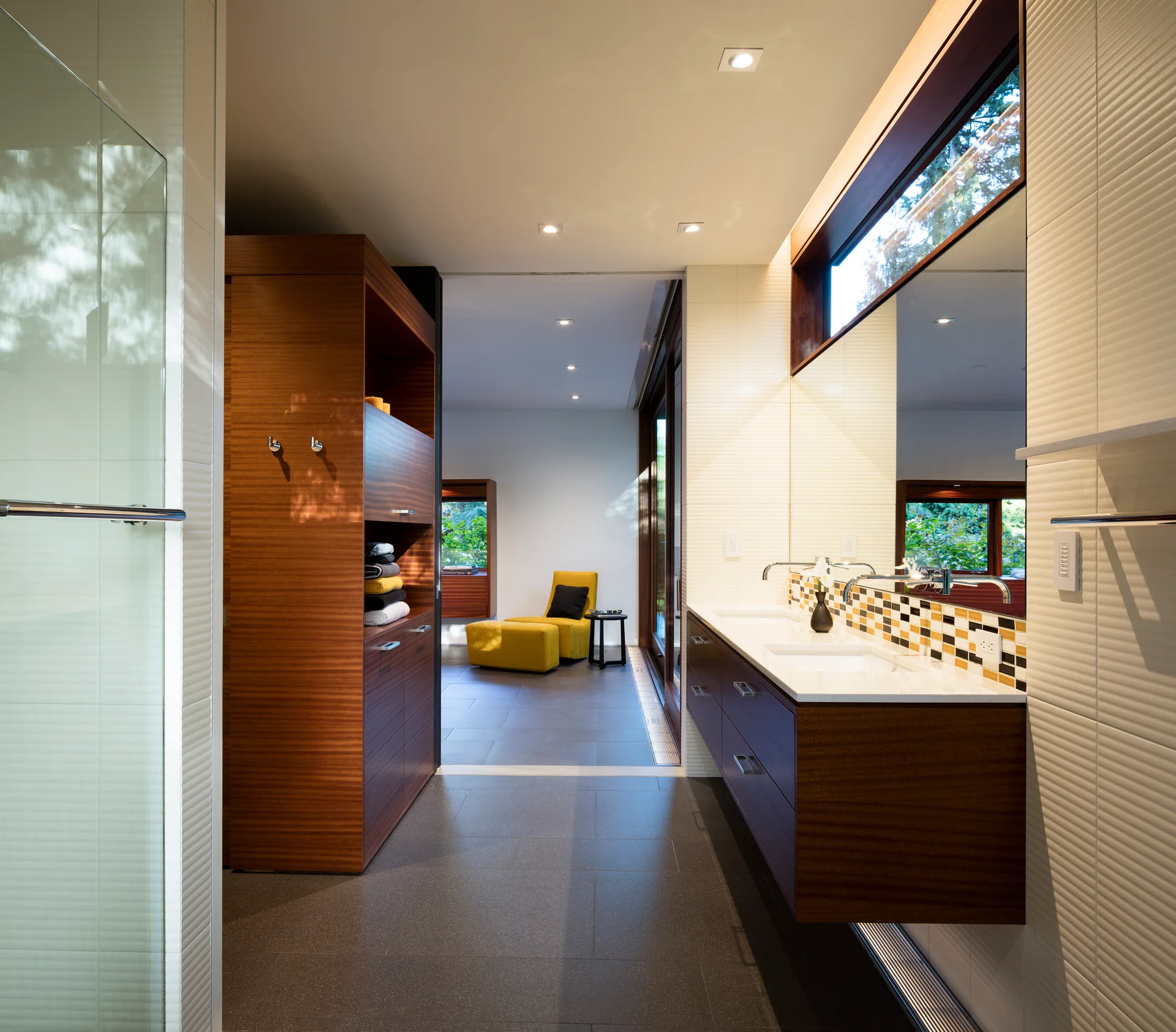 A modern bathroom features a dark wood floating dual vanity, a mosaic tile backsplash, a large mirror beneath a clerestory window, and grey floor tiles.
