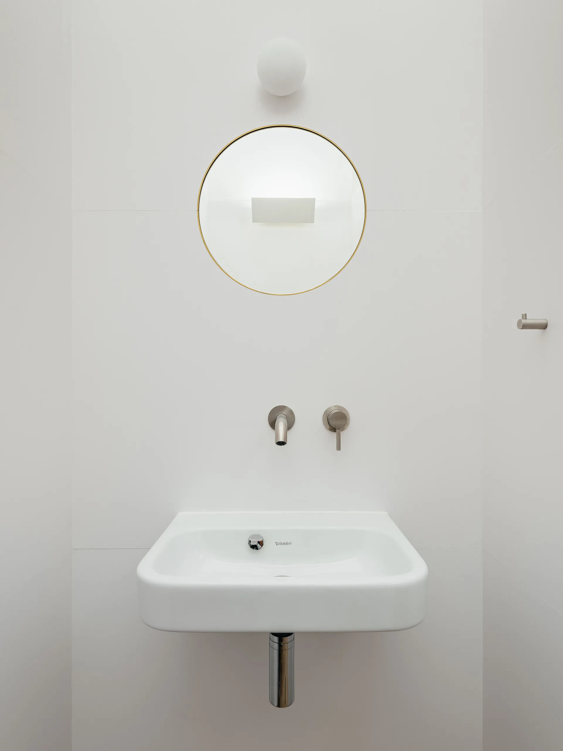 A wall-mounted white ceramic sink is paired with brushed metal faucet fixtures, a circular gold-trimmed mirror, and a small round wall sconce.