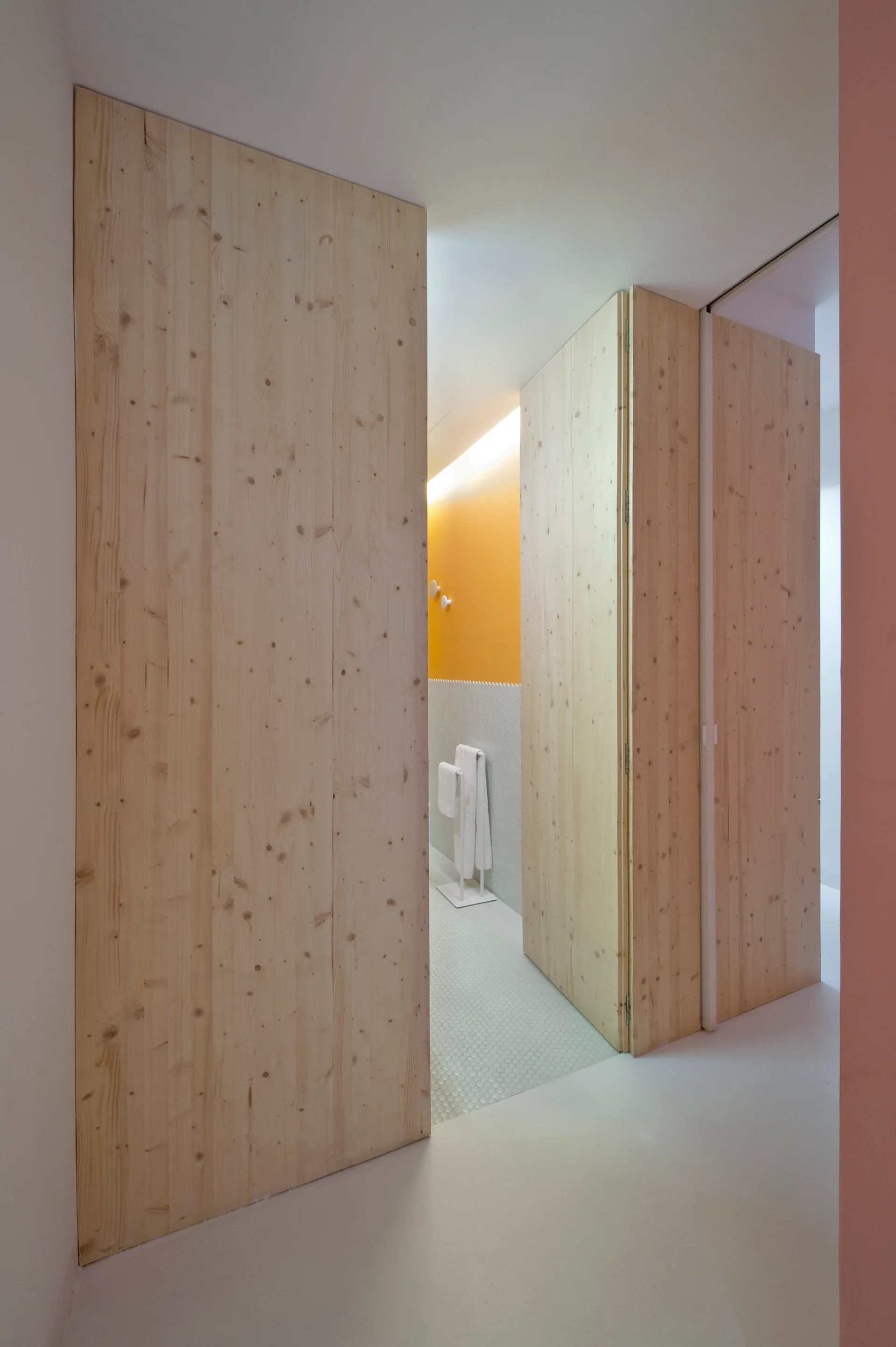 Large knotty pine flush doors stand open to reveal a bathroom with a bright yellow accent wall, white hexagonal tile flooring, and a white towel rack.