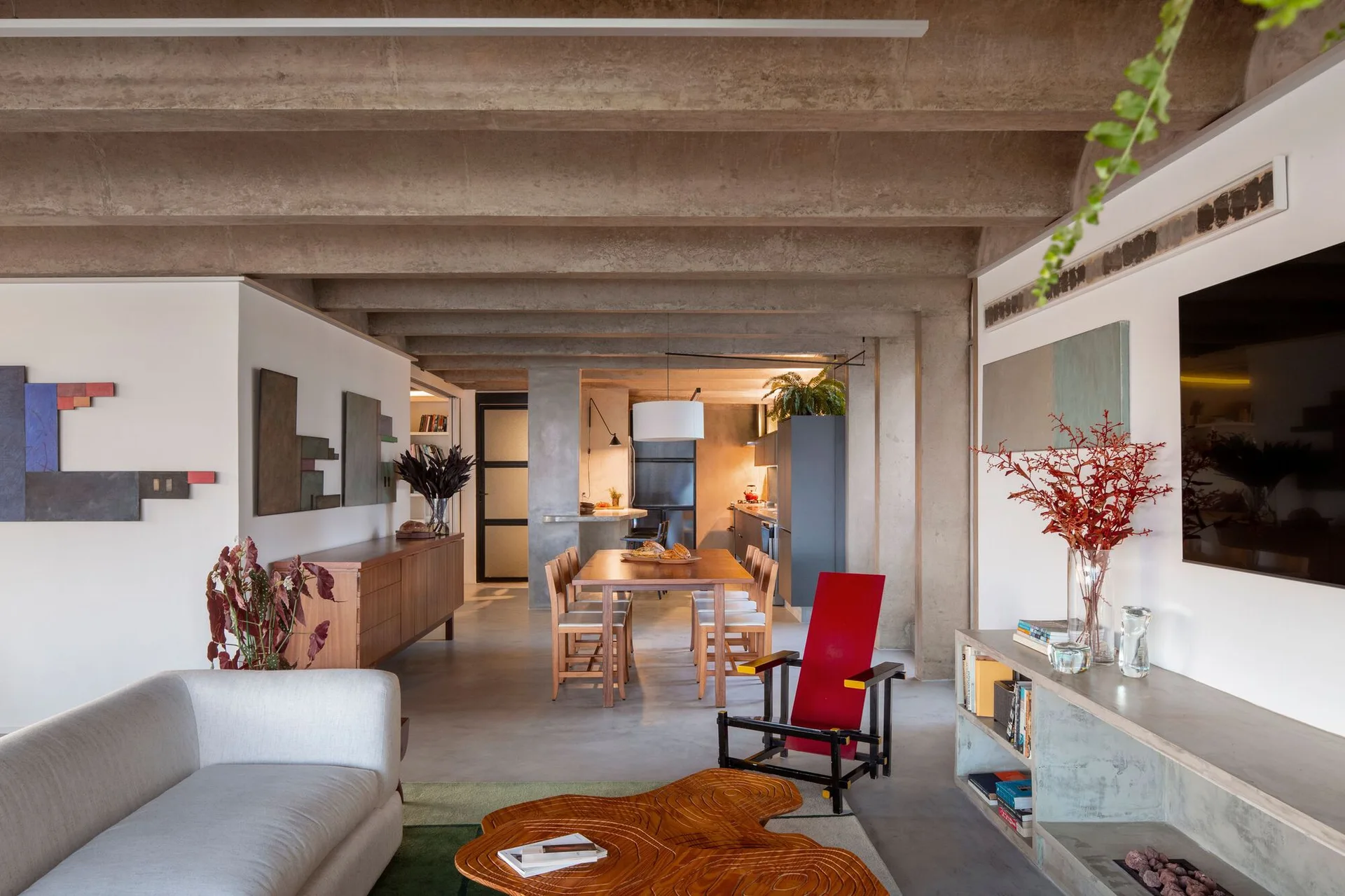 An open-plan living room features exposed concrete ceiling beams, a gray sofa, an organic wood coffee table, and a wooden dining table in the background.