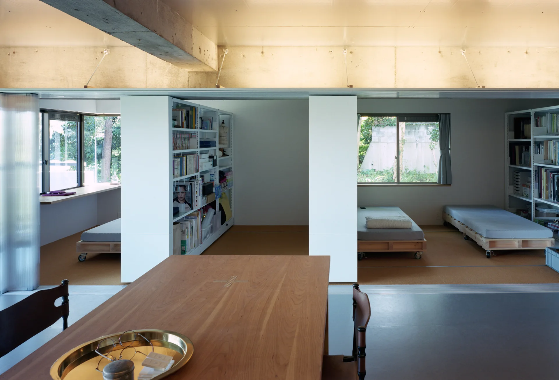 A minimalist multi-functional room featuring a wooden dining table in the foreground and rolling platform beds divided by white walls.