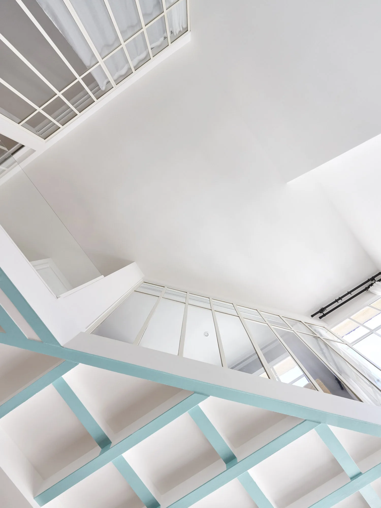 An upward low-angle view of a white staircase featuring light teal geometric wooden railings, glass grid partitions, and white ceilings.