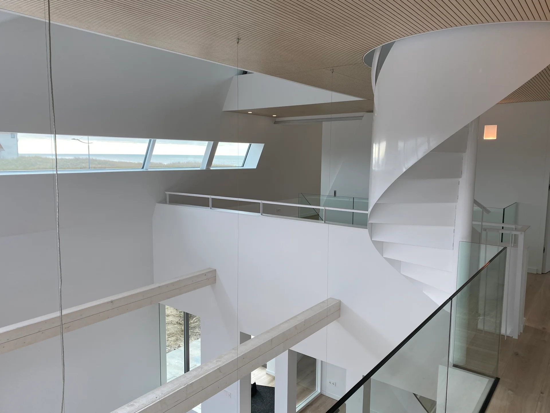 A minimalist split-level interior featuring a white spiral staircase, a light wood slatted ceiling, glass railings, and a horizontal window showing the ocean.