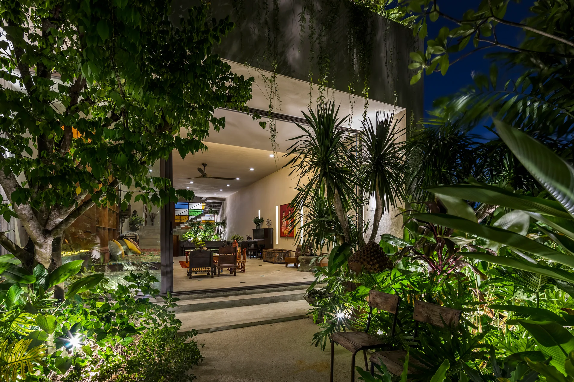 An open-concept living room with warm lighting transitions seamlessly into a lush, tropical garden at night through wide sliding doors.