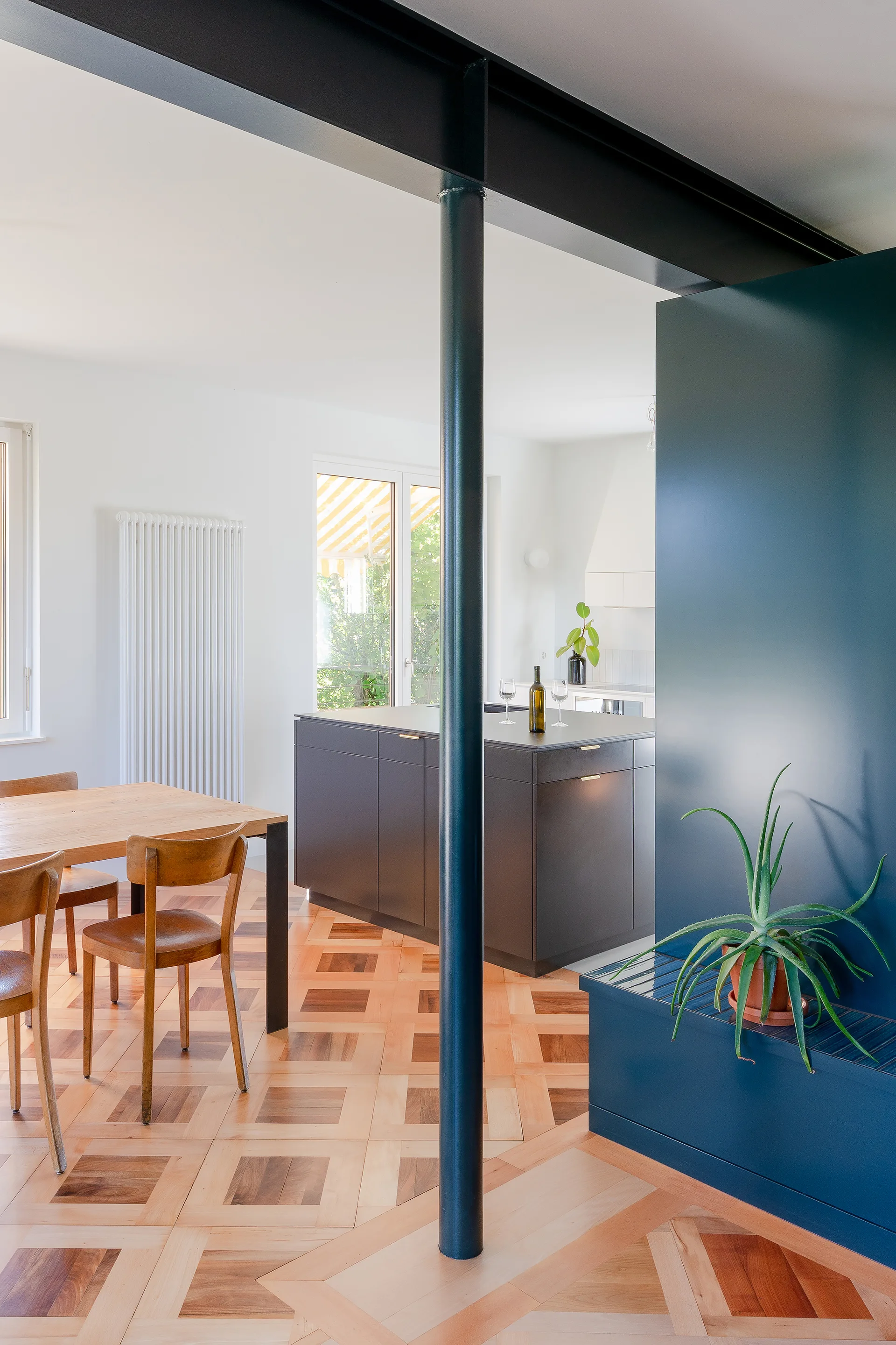 An open-plan kitchen and dining area features a dark grey island, wooden table and chairs, a teal partition wall, and patterned parquetry flooring.