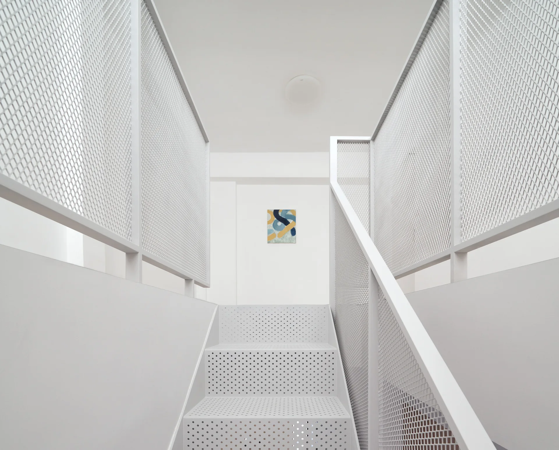 A minimalist white staircase features perforated metal treads and expanded mesh safety railings leading to a white landing.
