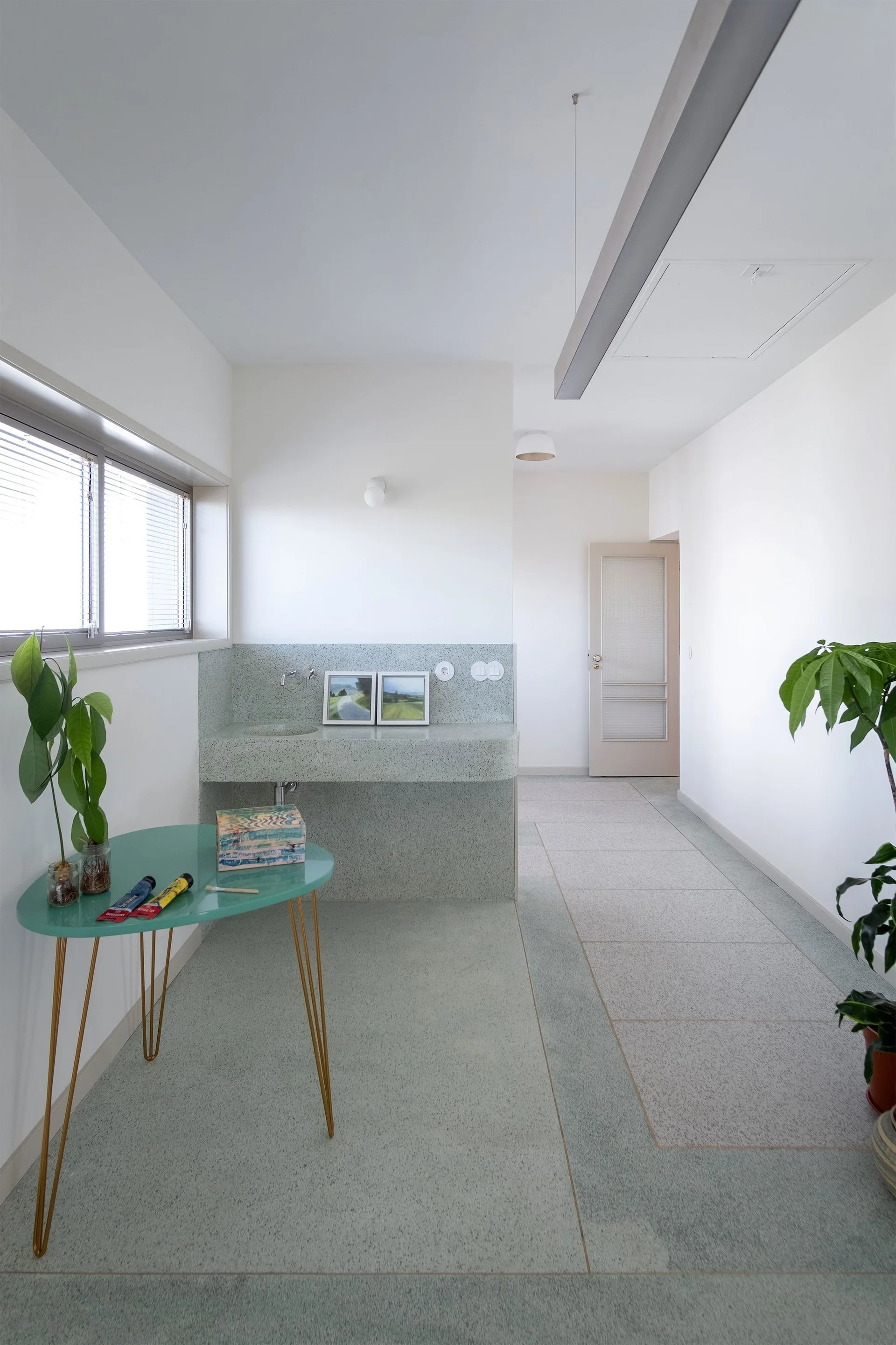 A minimalist hallway features a mint green terrazzo sink console, speckled floor tiles, a teal side table with plants, and plain white walls.
