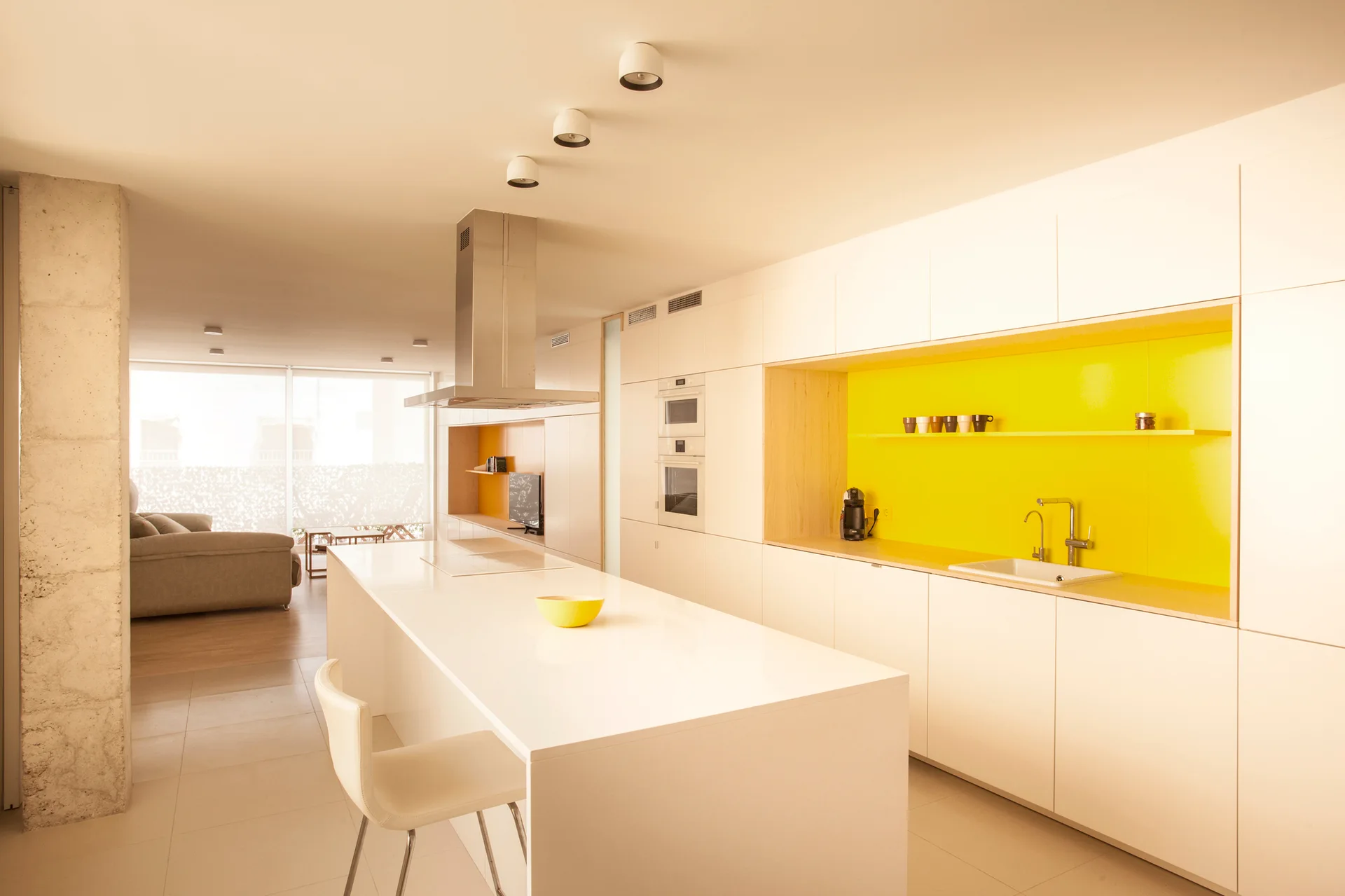 A modern minimalist kitchen features seamless white cabinetry, a large central island, and a recessed bright yellow backsplash shelf.