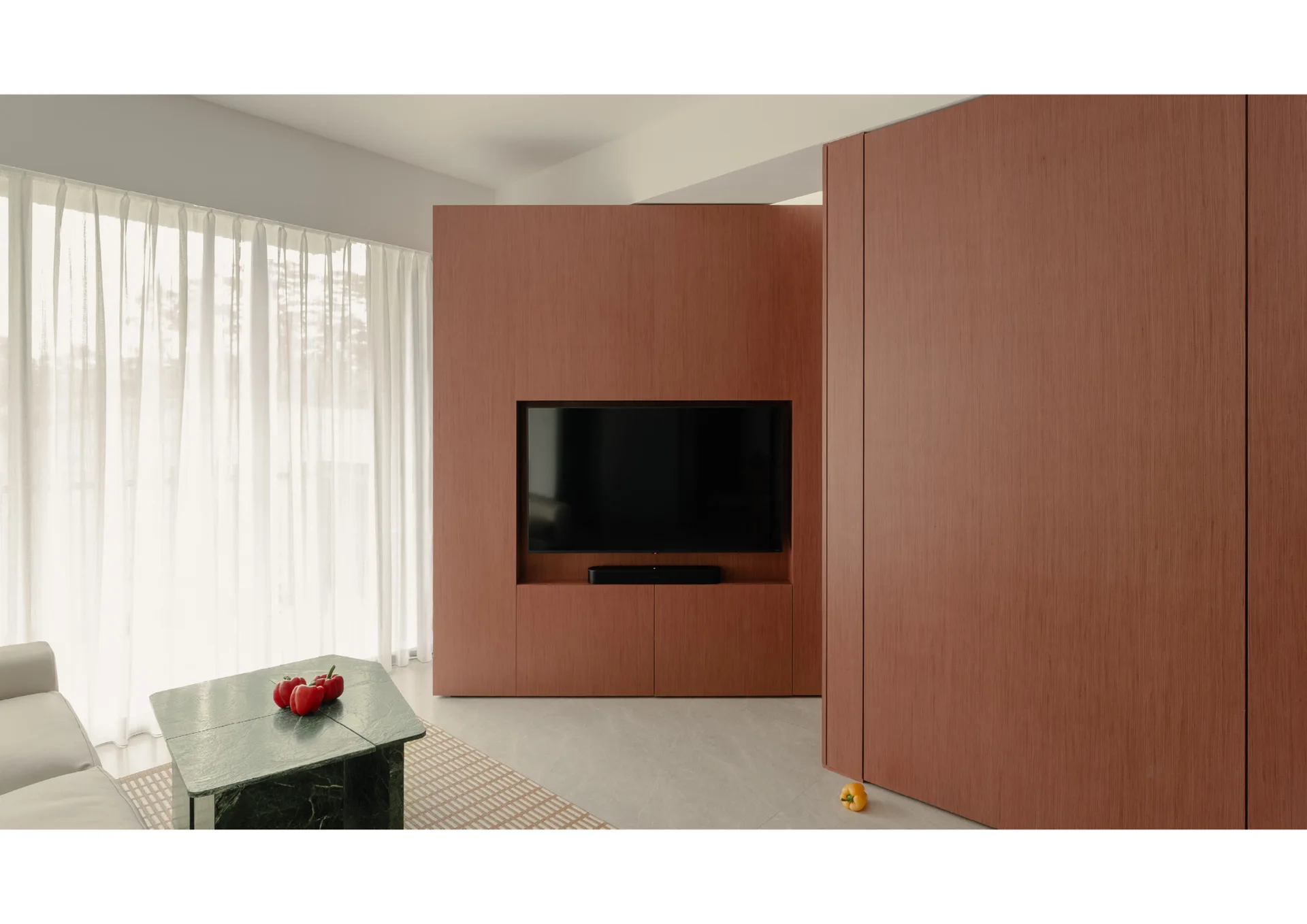 A minimalist living room featuring a television recessed in a warm wood paneled wall next to a green marble coffee table and sheer curtains.