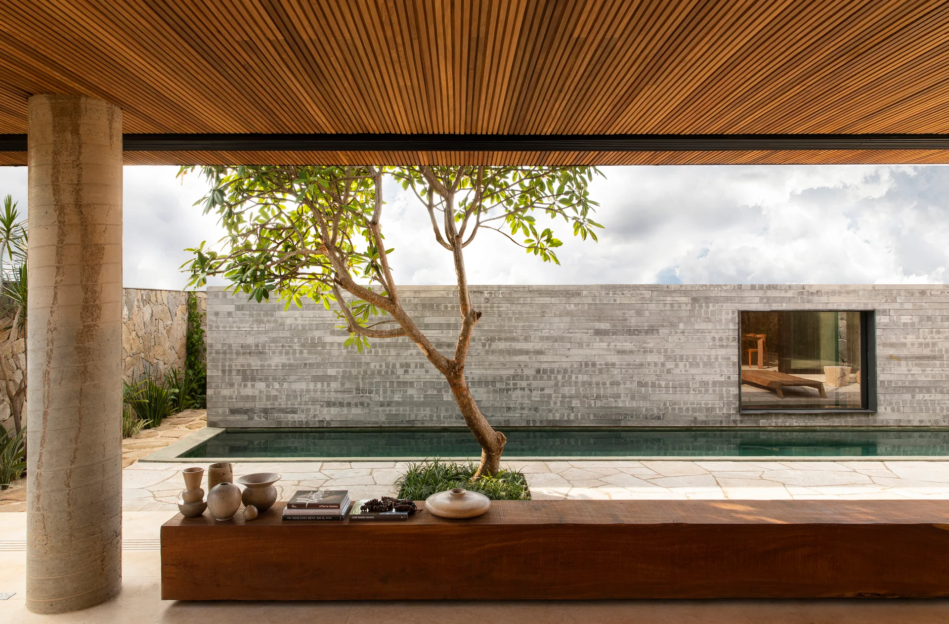 A covered outdoor patio features a long wood bench, a concrete column, a central tree, and a narrow lap pool in front of a grey stone wall.