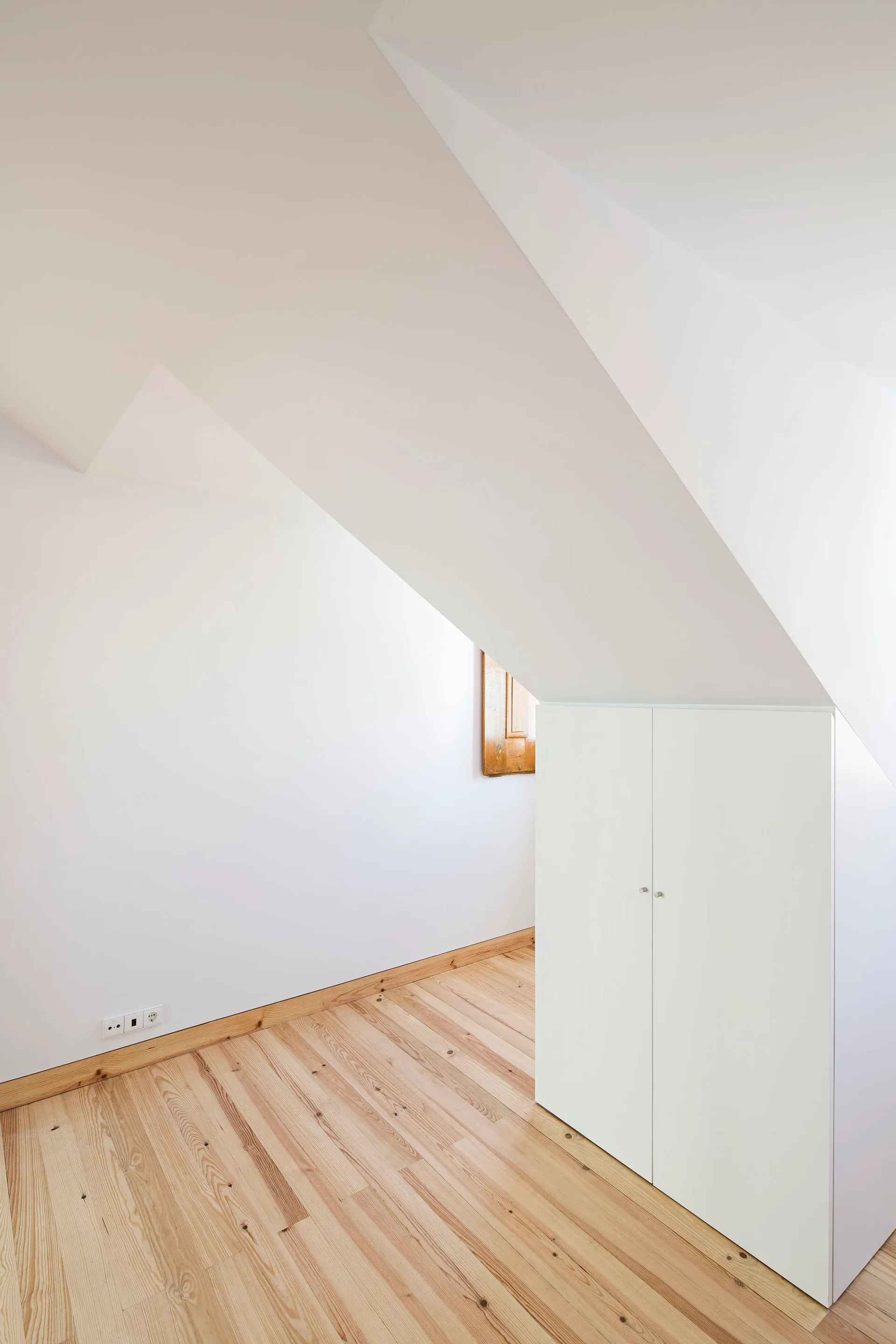 An attic interior with sloped white walls, light wood floors, and a simple two-door built-in white cabinet tucked under the roof angle.