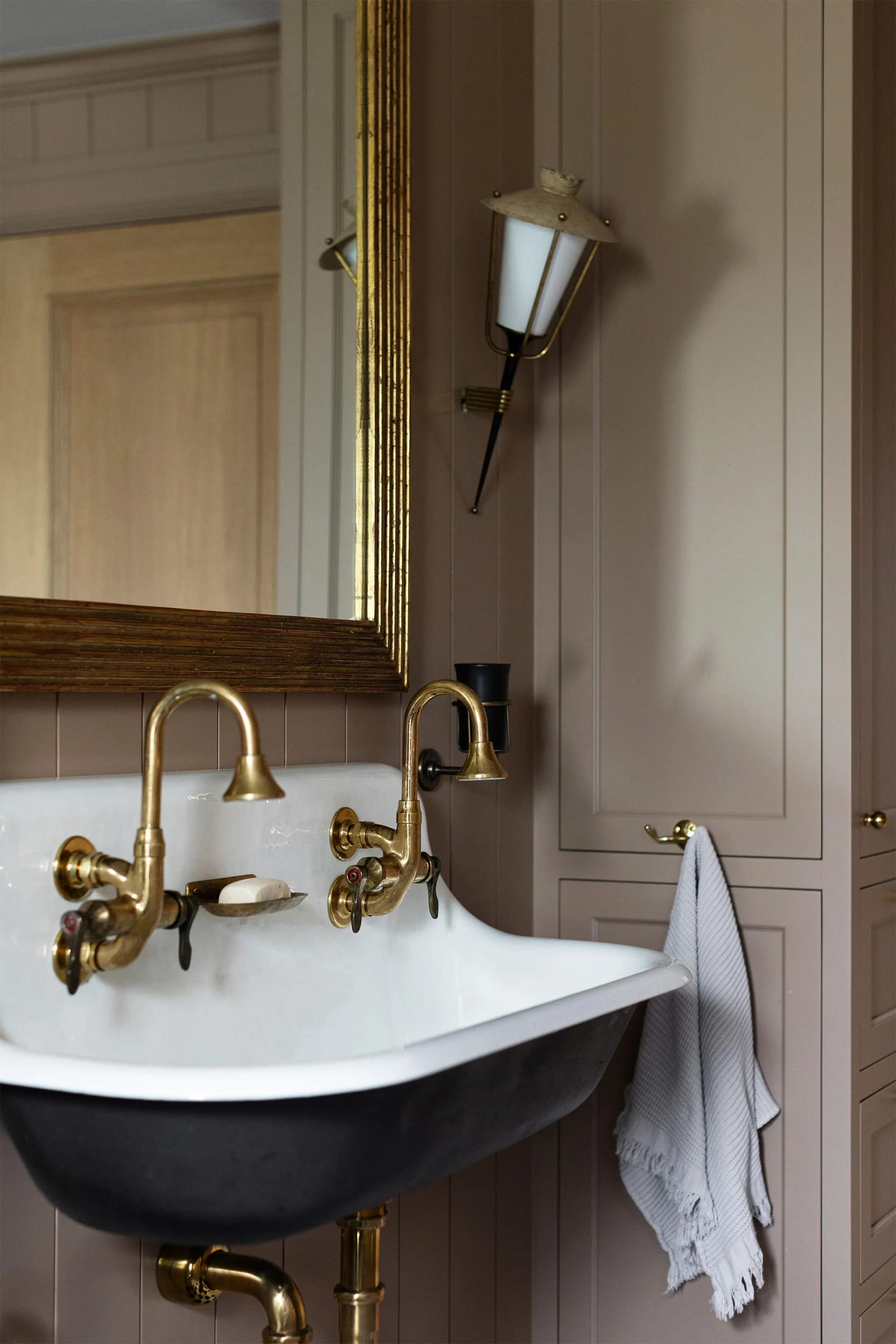 A wall-mounted white porcelain sink with two vintage brass faucets is set against taupe-painted wood wall paneling in a traditional bathroom.