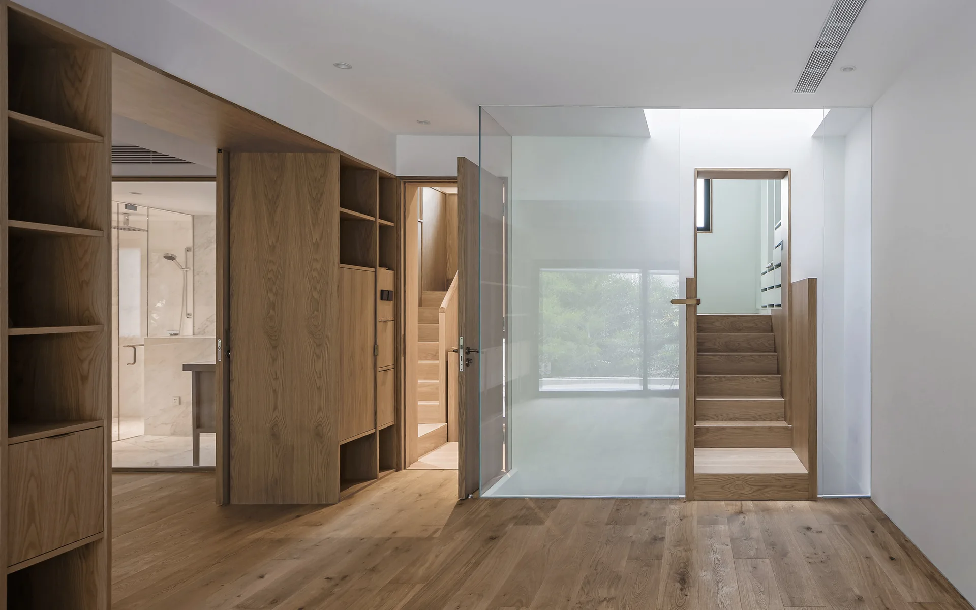 Minimalist interior hallway with a wooden staircase behind a glass partition, custom oak cabinetry, and matte wood flooring.