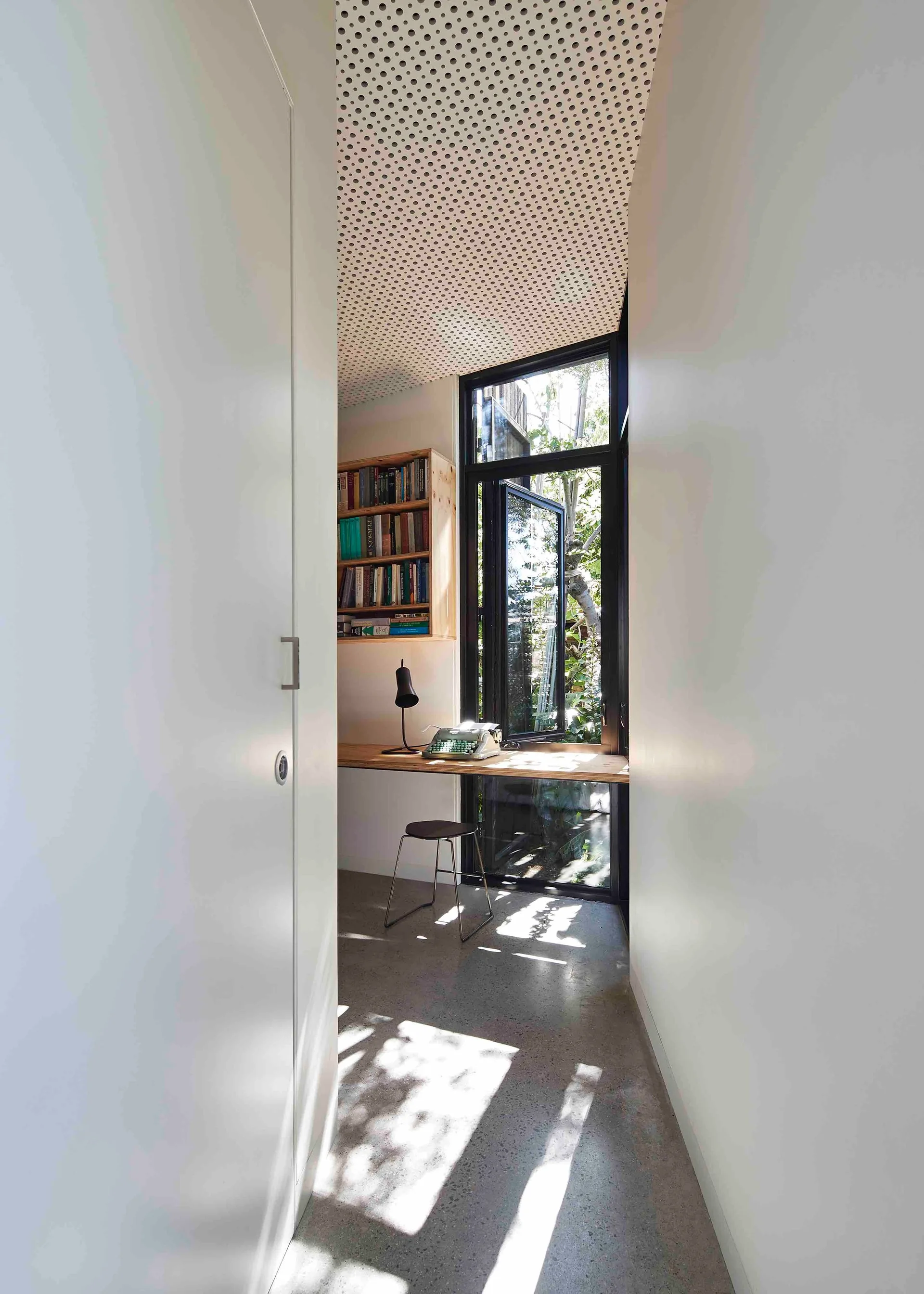 A narrow minimalist home office features a long wooden floating desk, a wall-mounted bookshelf, a simple stool, and a large black-framed window.