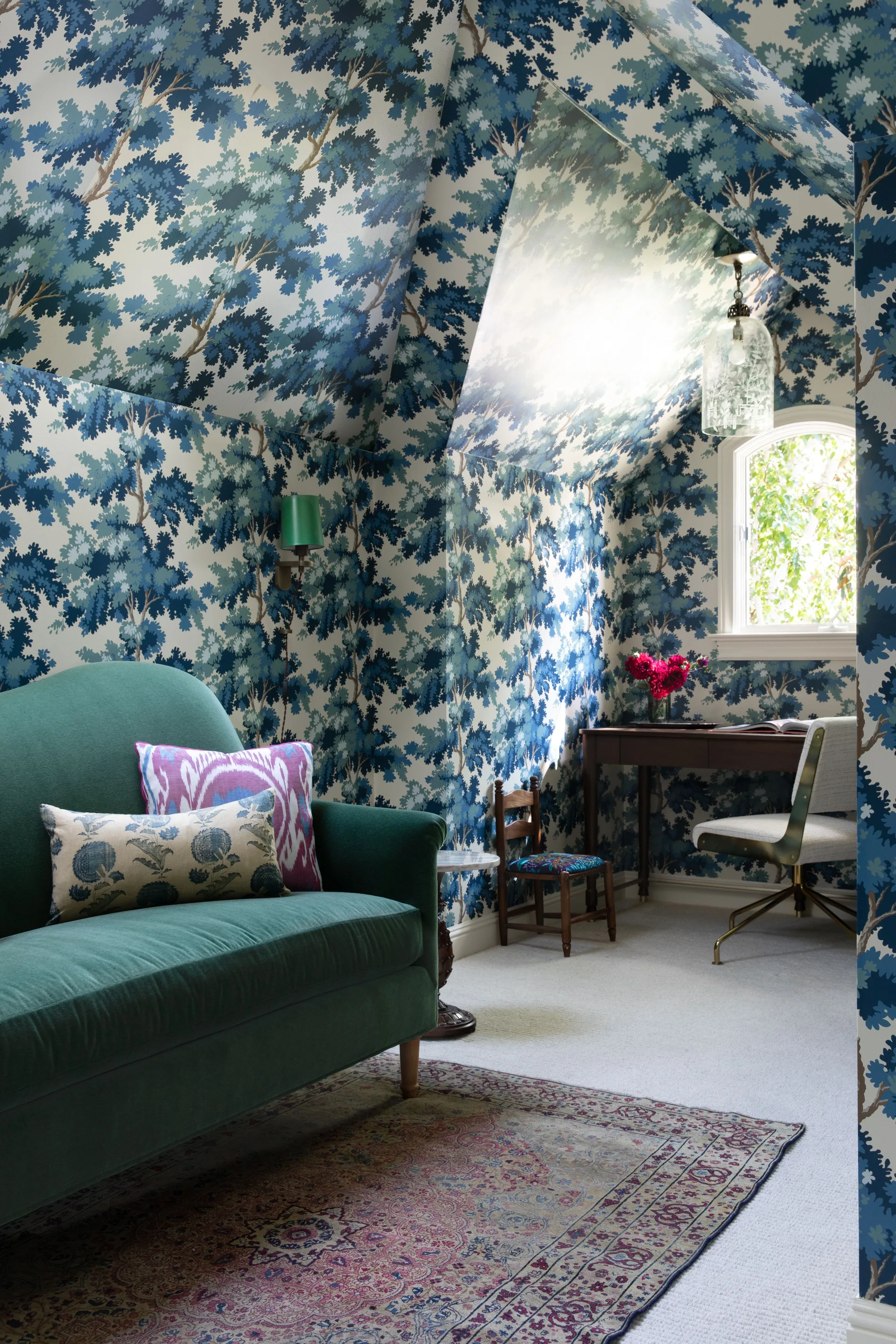 A home office with blue tree-print wallpaper covering the sloped ceiling, a green velvet sofa, and a wood desk near an arched window.