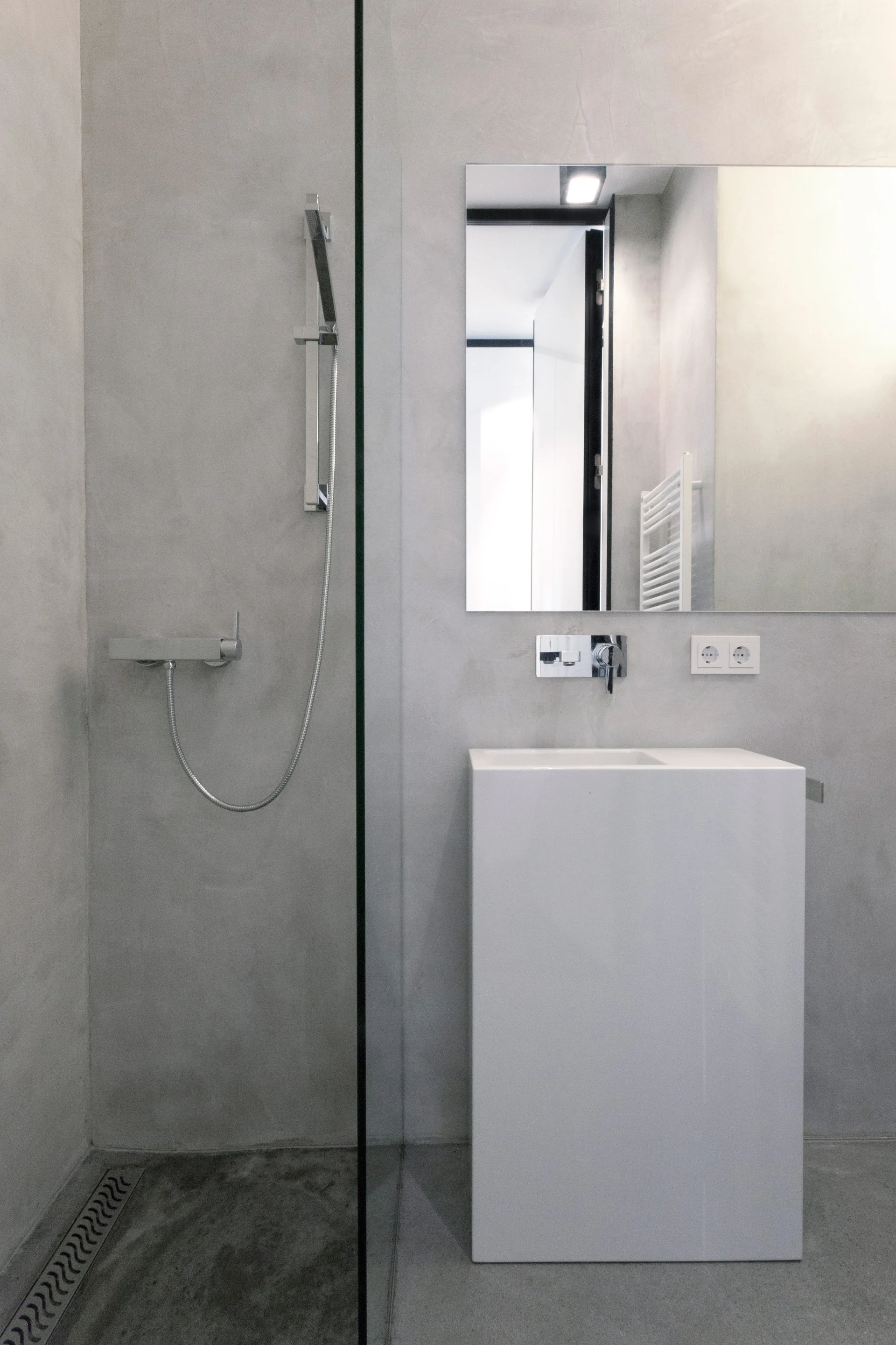 A minimalist bathroom with smooth grey concrete walls features a rectangular white pedestal sink, a frameless wall mirror, and a glass shower partition.