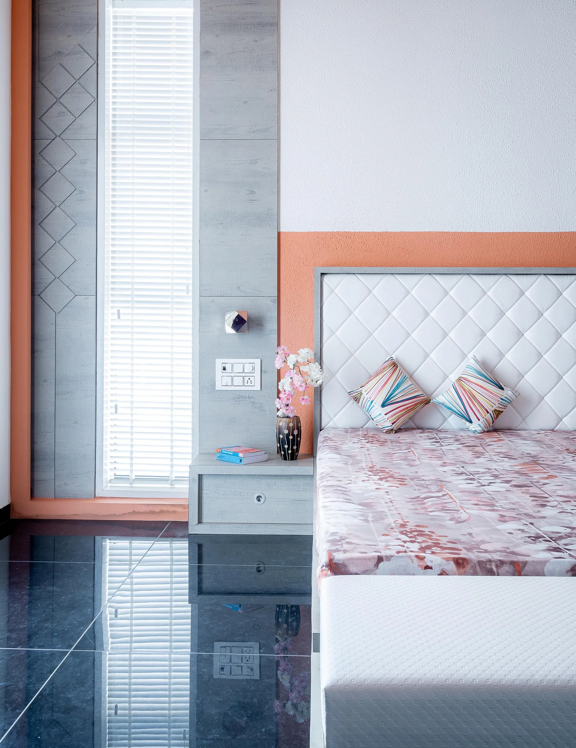 A modern bedroom corner featuring a quilted white headboard, floating grey nightstand, and a narrow window next to an orange wall accent.
