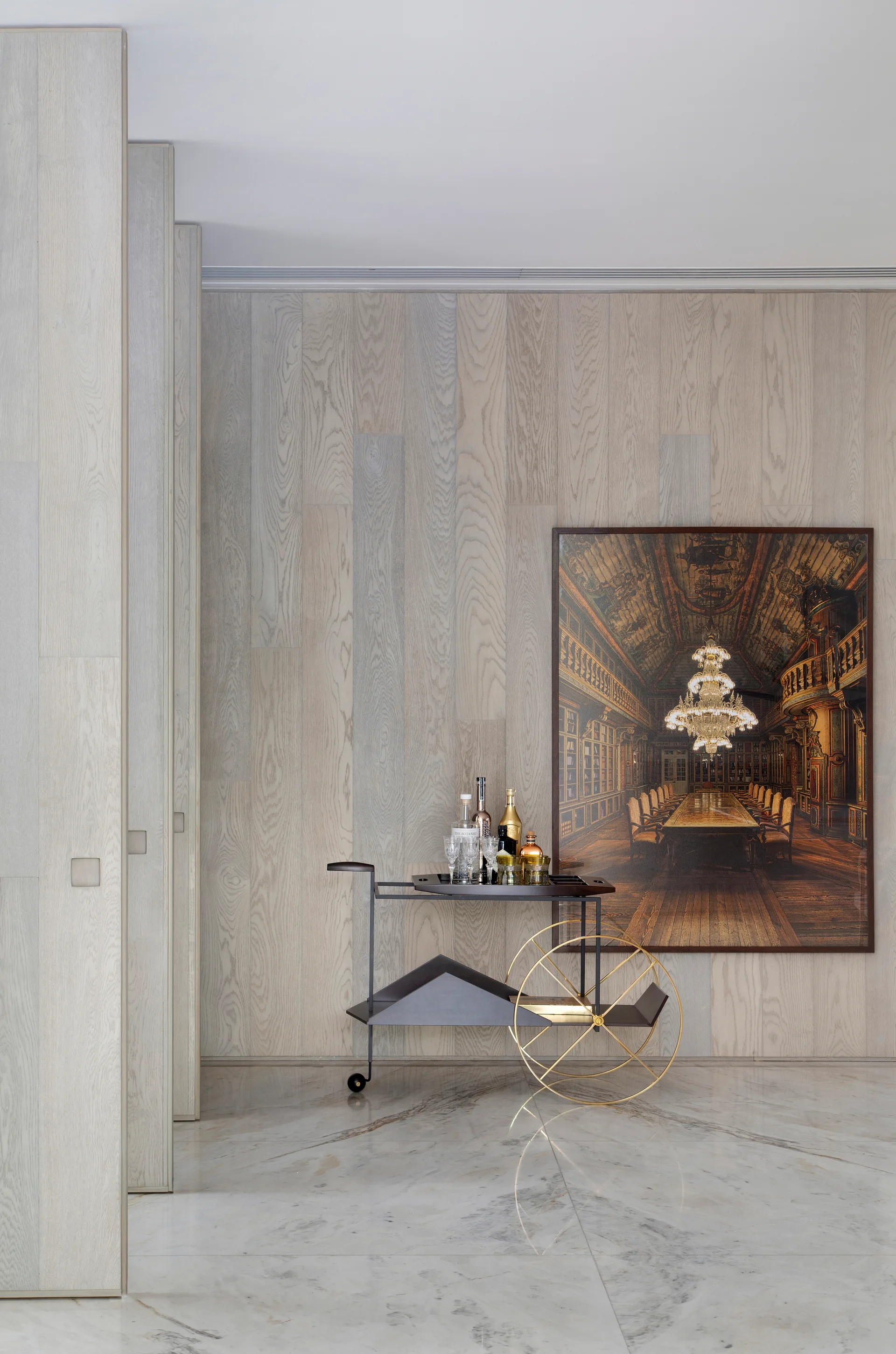 A modern black bar cart with large gold wheels stands on a polished marble floor in front of a light wood-paneled wall and large framed artwork.