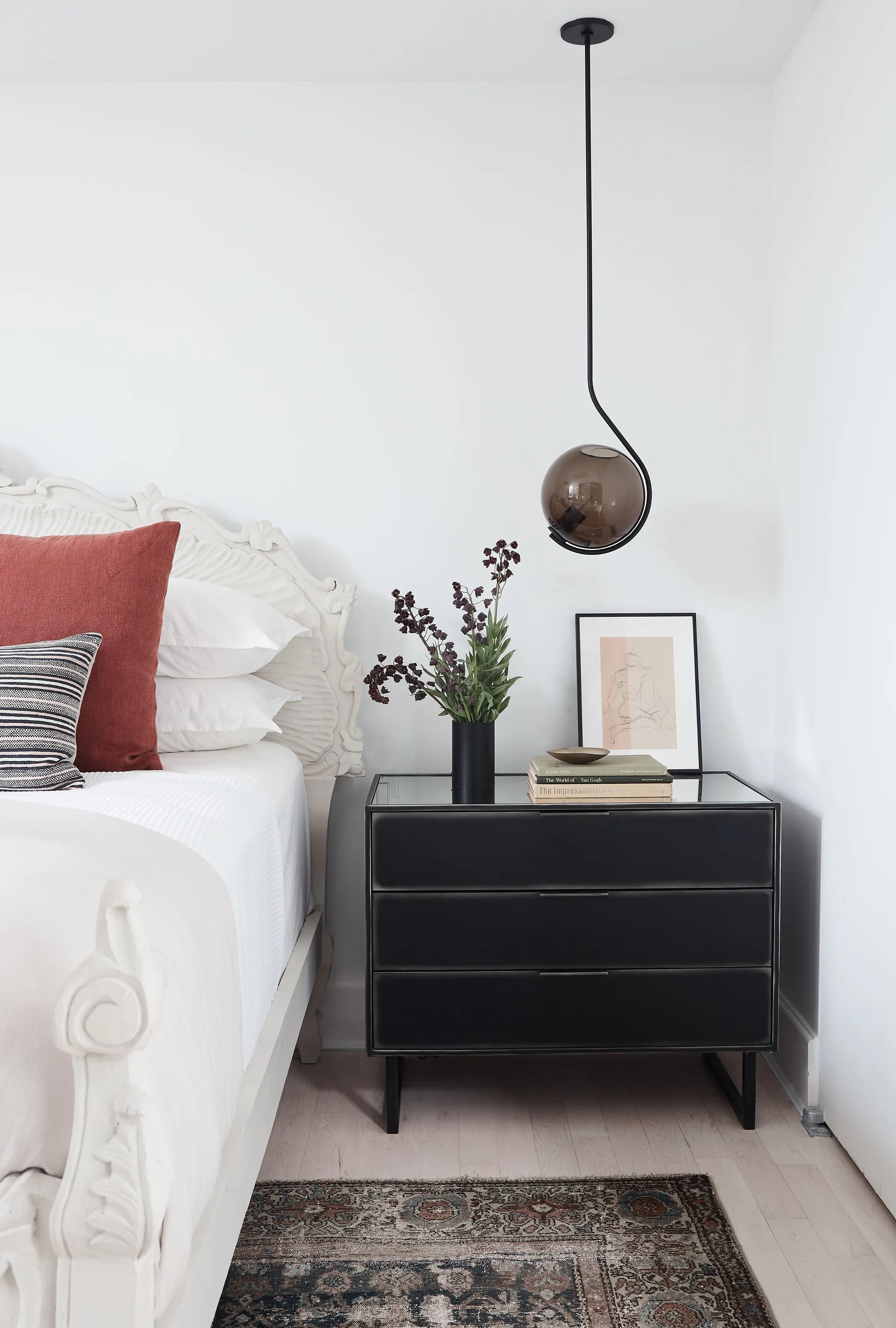 A bedroom featuring an ornate white carved wood headboard next to a three-drawer black nightstand with an amber glass pendant light hanging above.