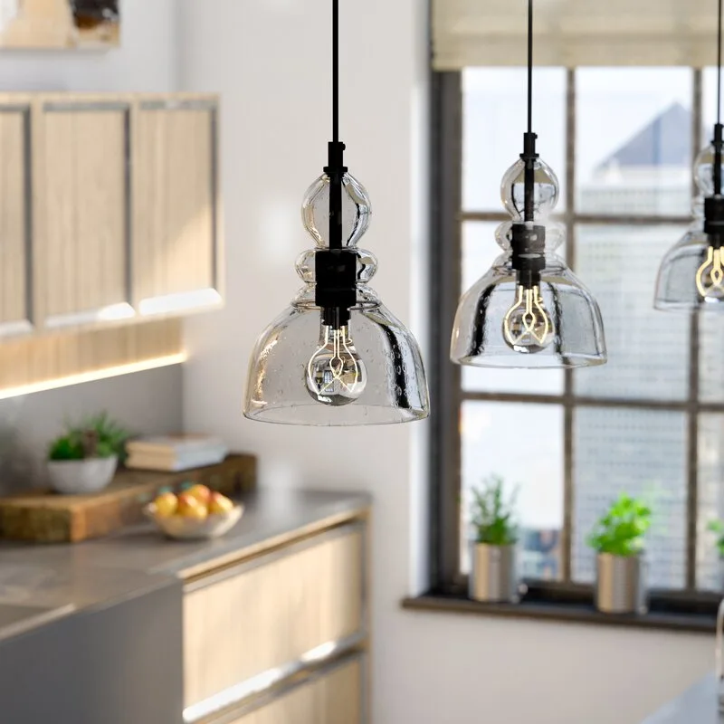 Three clear glass pendant lights with warm filament bulbs hanging above a grey kitchen counter in front of a black metal grid window.