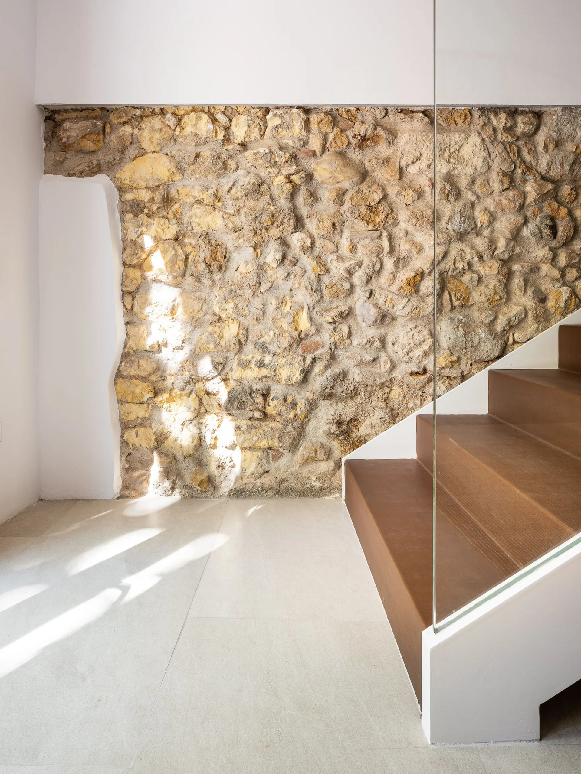 A modern staircase with brown wood treads and a glass railing next to a textured, exposed natural stone feature wall.