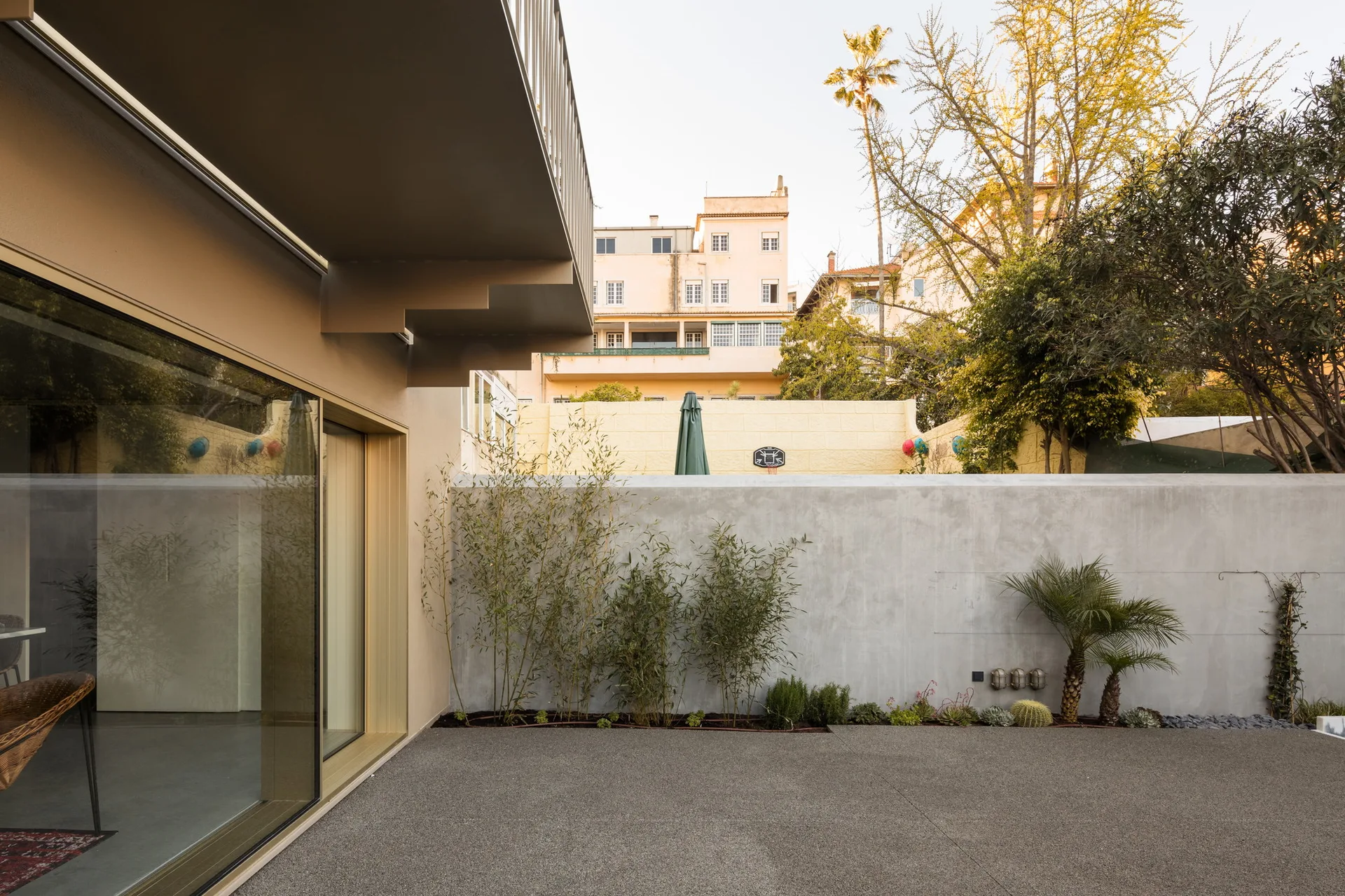 A minimalist outdoor courtyard featuring a high concrete privacy wall, textured gravel paving, and a garden border planted with slim bamboo.