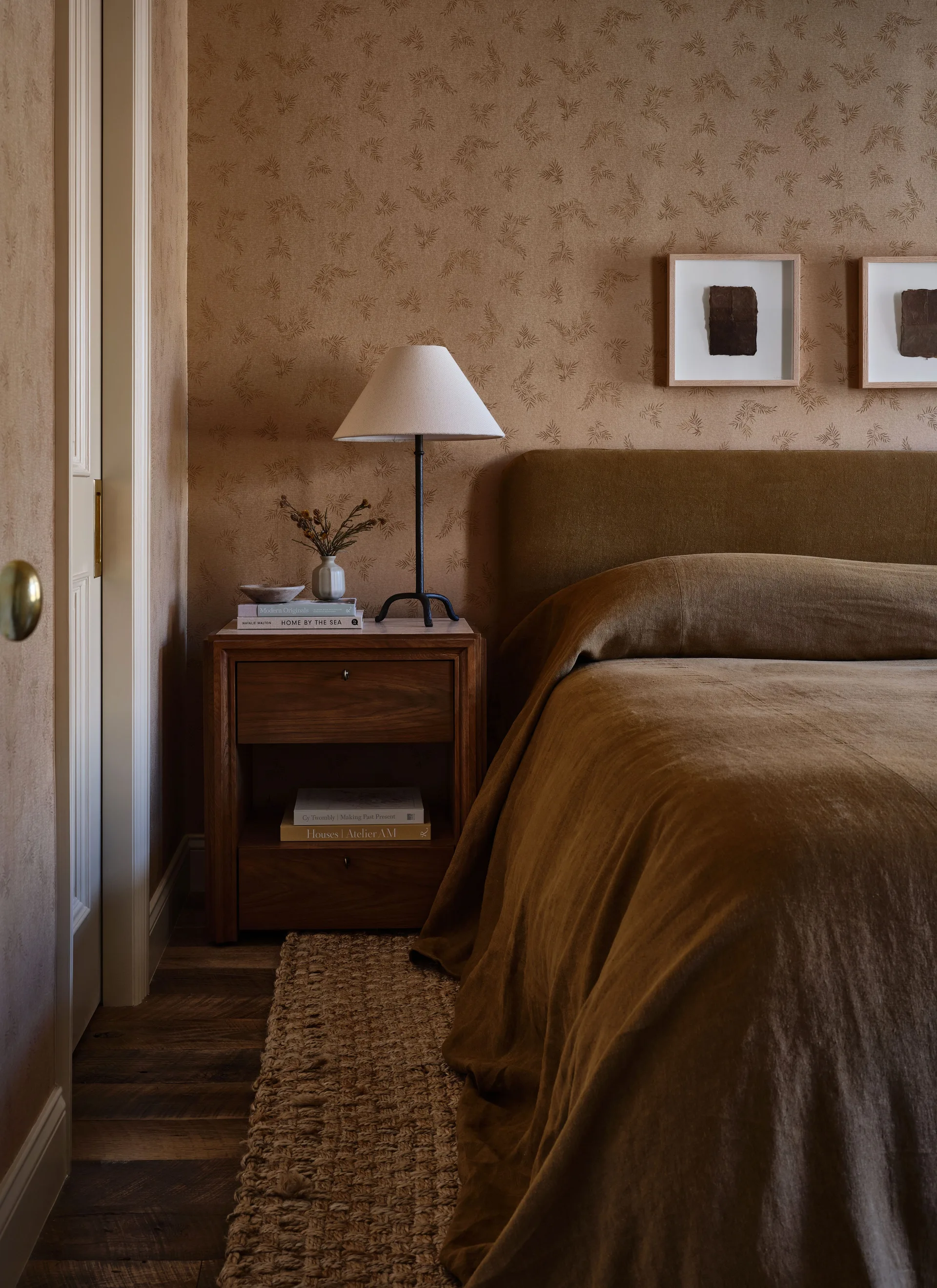 A bedroom features a bed with deep brown linens next to a wooden nightstand with a table lamp against tan botanical wallpaper.